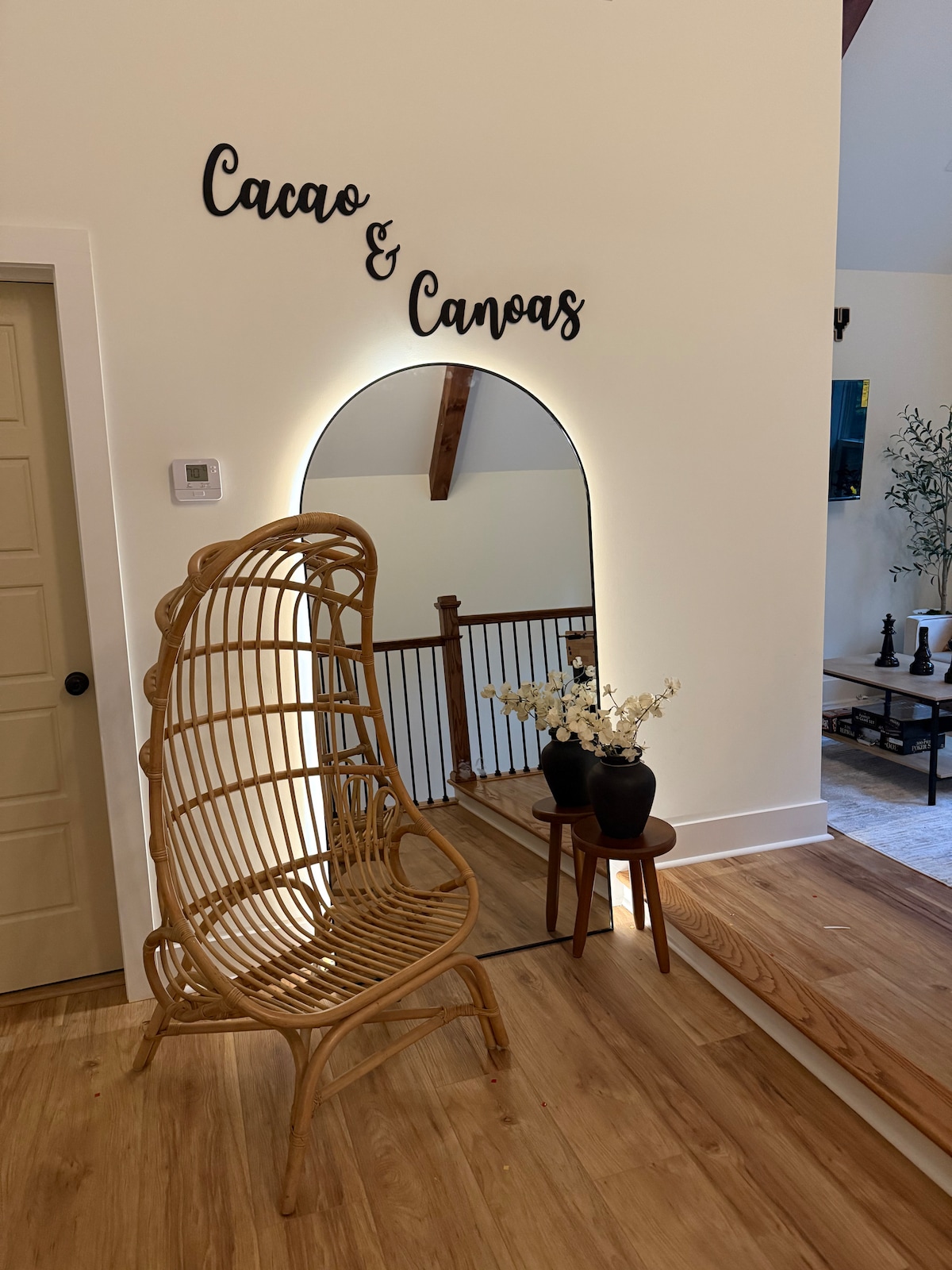 A welcoming entryway features a decorative sign reading 'Cacao & Canoas'. A uniquely designed wicker chair adds character, with a potted plant resting atop a small table nearby. Soft lighting enhances the space against pale walls and wooden flooring.
