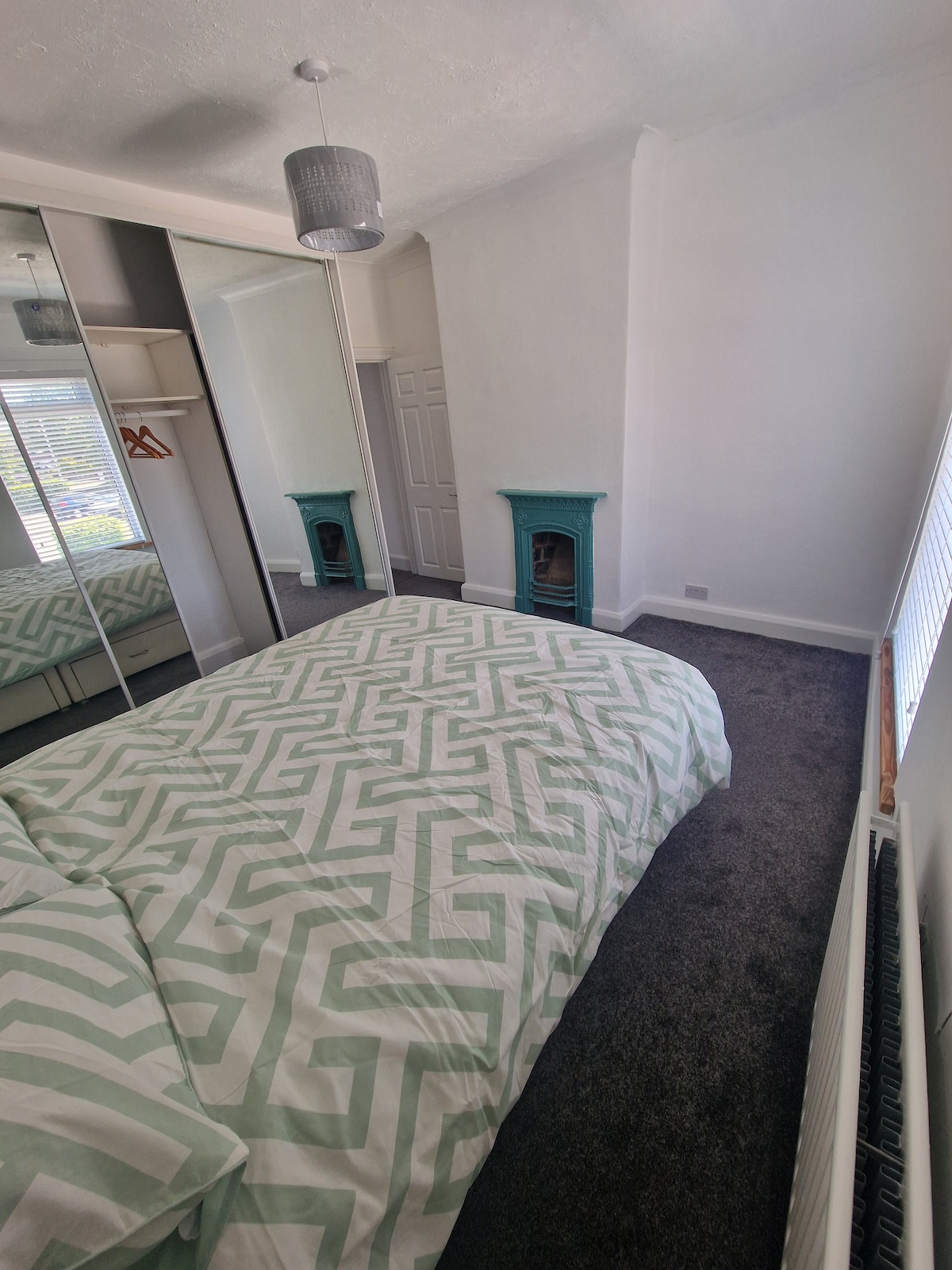 A bright bedroom features a spacious bed adorned with a patterned duvet cover. A mirrored wardrobe provides ample storage, reflecting natural light. A decorative fireplace adds character, accompanied by a radiator along the wall. A window allows soft light to illuminate the room.