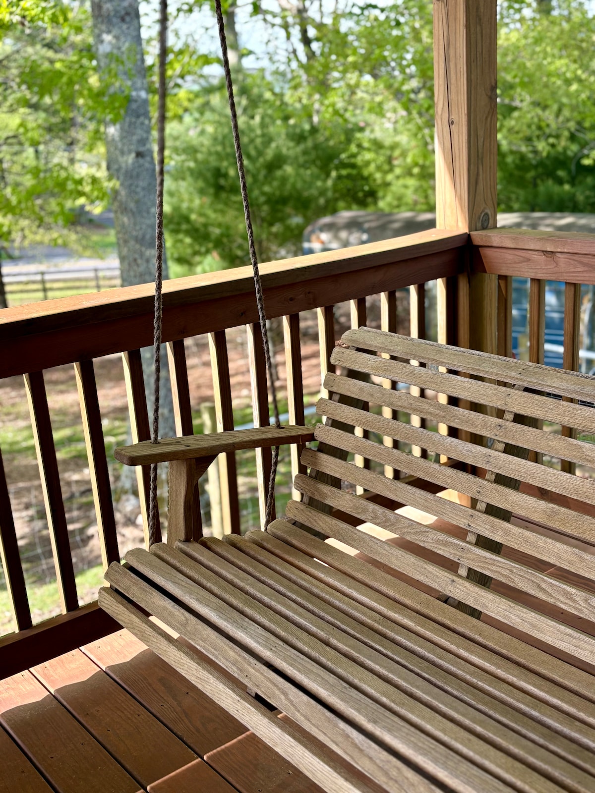 A wooden swing bench is positioned on a covered porch, inviting moments of relaxation. The swing features slatted seating and is suspended by sturdy ropes, offering views of the surrounding greenery and peaceful landscape.