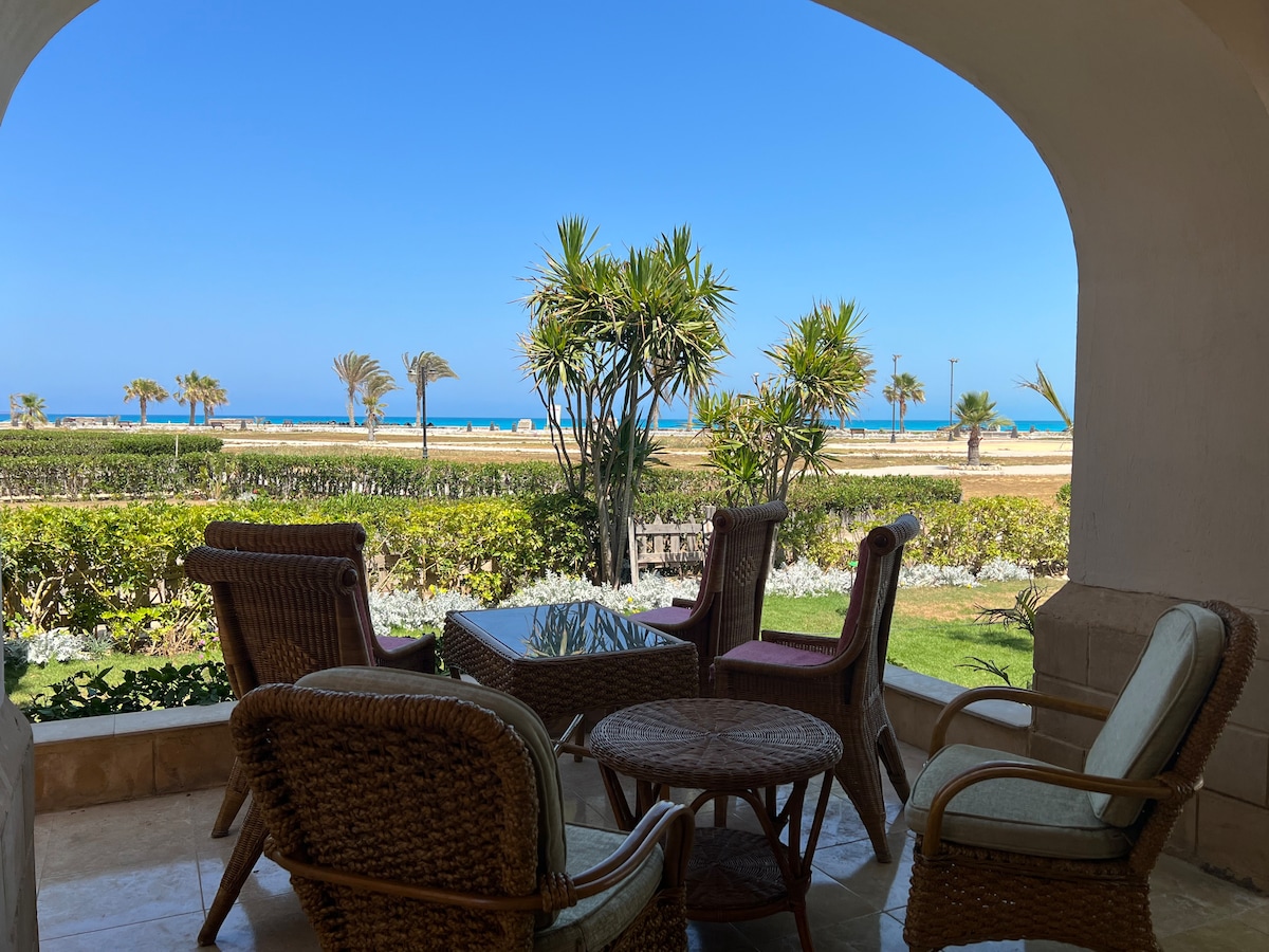 A shaded outdoor seating area is presented, featuring wicker chairs surrounding a small round table. Lush greenery and flowering plants are visible nearby, while the glistening beach and ocean stretch beyond, framed by clear blue skies.