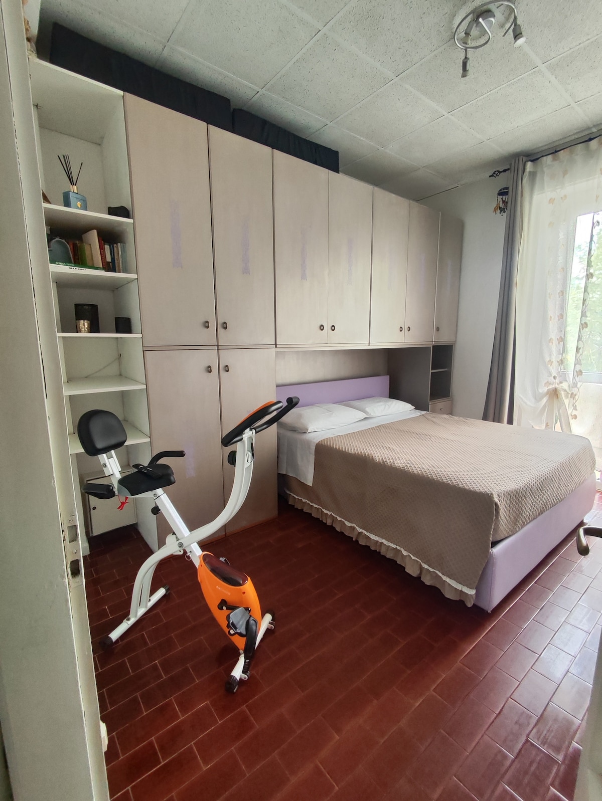 The bright bedroom features a spacious bed covered in a neutral-toned blanket, complemented by built-in cabinetry along one wall. A stationary bike is positioned nearby, contributing to the room's fitness-friendly atmosphere. Natural light enters through a nearby window adorned with light curtains.