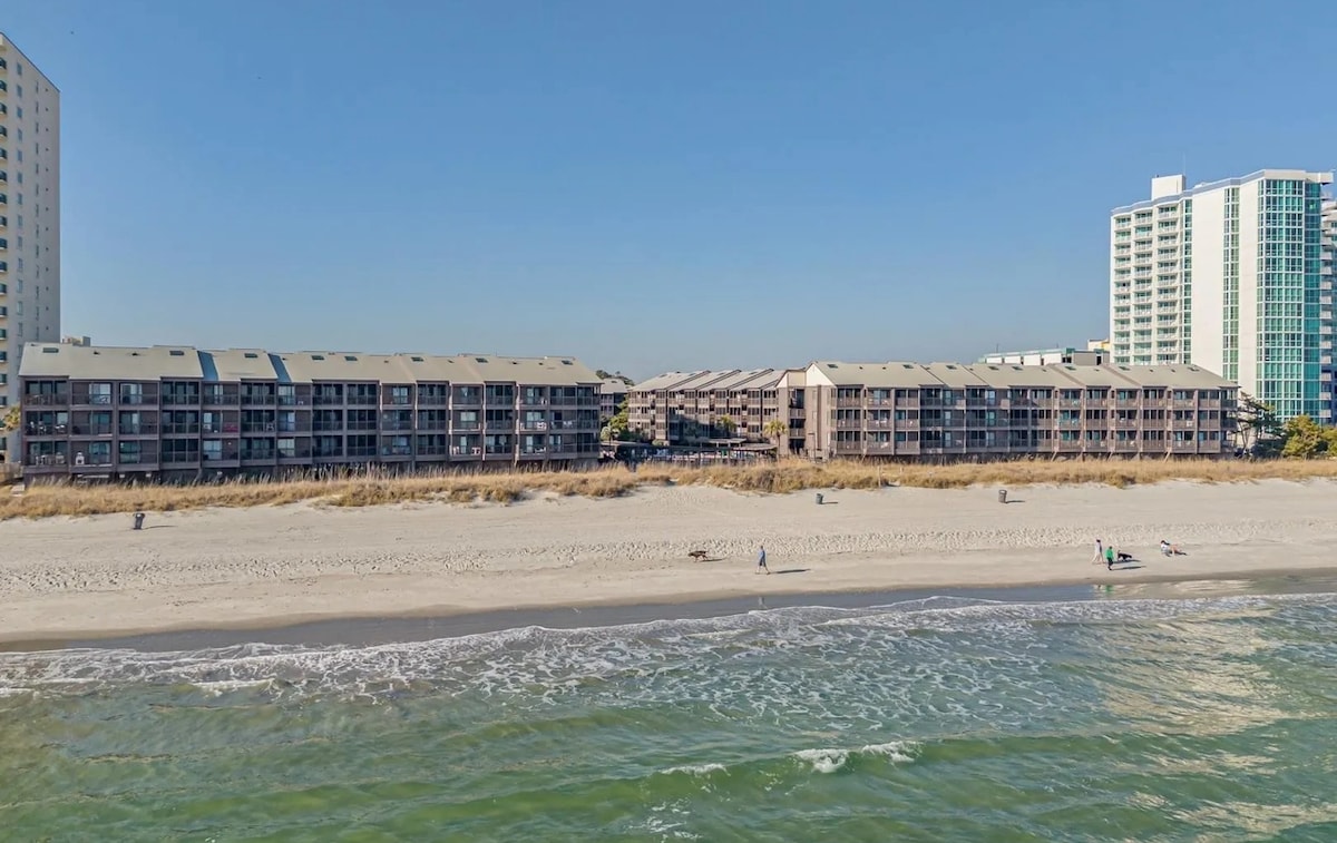 An expansive view captures a series of beachfront condos along a sandy shore, bordered by gentle waves. The surrounding scene features several people enjoying the beach, with tall structures visible in the background against a clear blue sky.