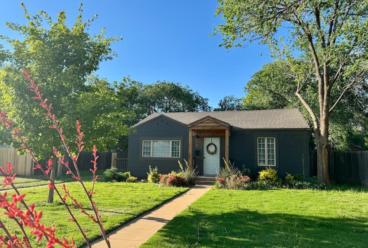 A charming updated house is framed by green grass and landscaped gardens. The entrance features a welcoming front porch with a wreath, and large windows allow natural light to fill the interior. Trees provide shade in the surrounding area.