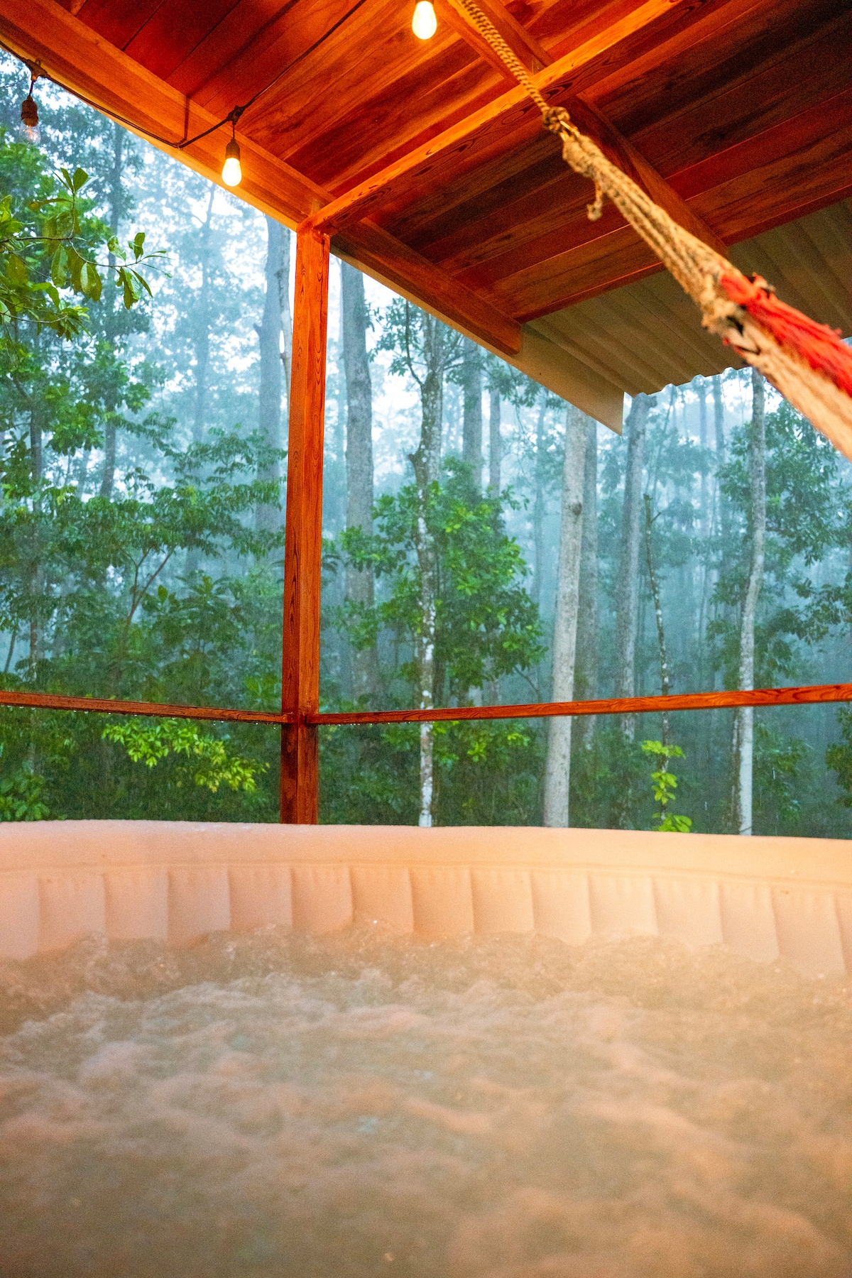 A cozy outdoor jacuzzi is seen, surrounded by a wooden deck and enveloped by lush trees. Soft lighting hangs above, creating an inviting ambiance. The misty forest backdrop enhances the serene atmosphere for relaxation.