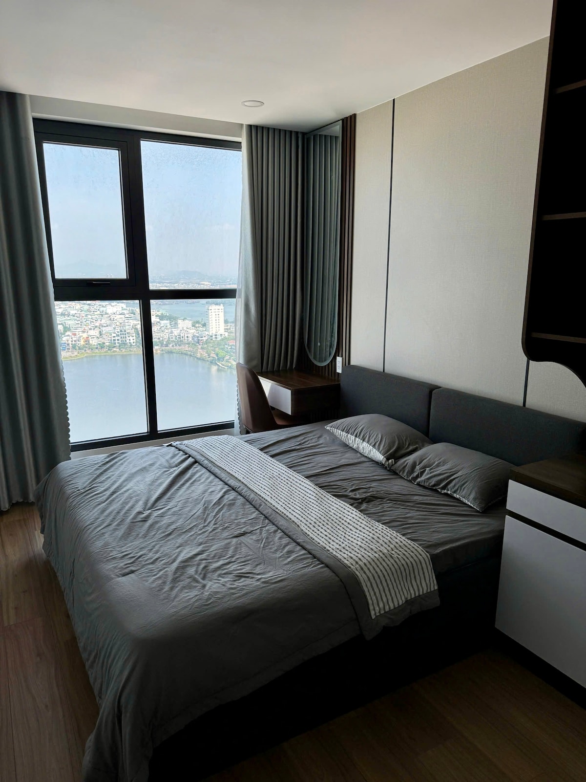 A cozy sleeping area features a spacious bed dressed in neutral linens. Large windows provide views of the cityscape and a nearby body of water. Soft drapes frame the windows, and a minimalist nightstand complements the modern decor.
