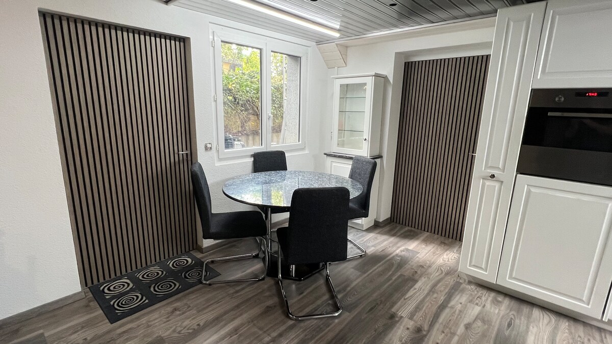 A sleek dining area is presented with a round glass table surrounded by four black upholstered chairs. Natural light enters through two windows, illuminating the space. The walls feature vertical wooden slats, contributing to a modern aesthetic, while a patterned rug adds texture to the floor.