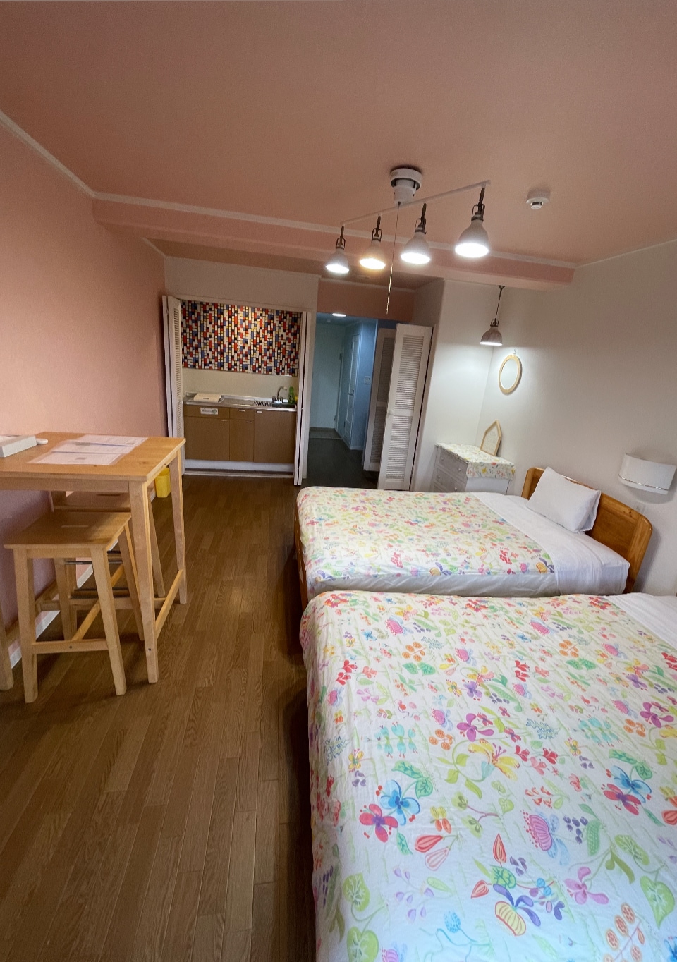 A room featuring two spacious beds adorned with floral patterned blankets. A small wooden table and stools are positioned beside the beds. The walls are painted in a soft pink, and a kitchenette is visible in the background.