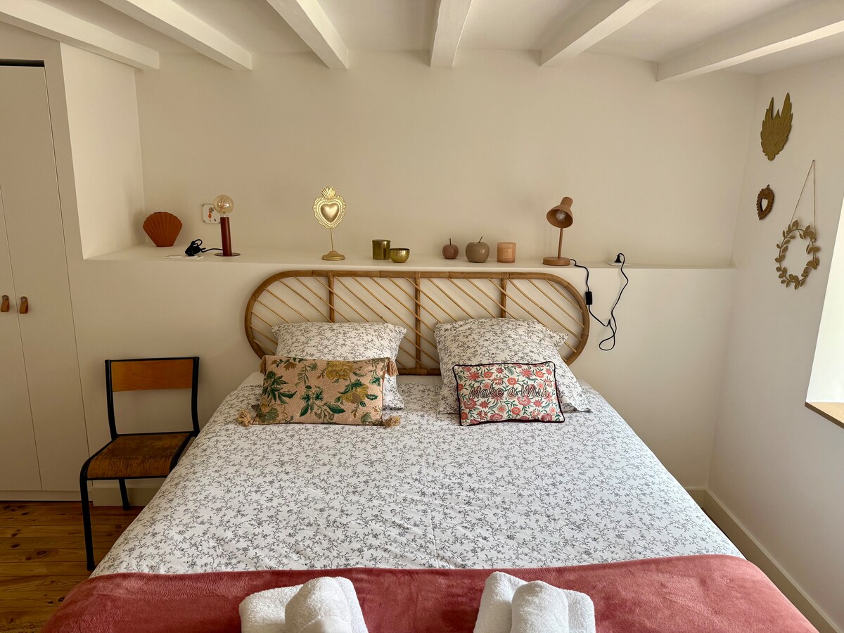 A cozy bedroom features a spacious bed with patterned linens and decorative pillows. Accents include a wooden headboard and various decorative items on a shelf above the bed. Natural light enters through a nearby window, enhancing the bright and airy atmosphere.