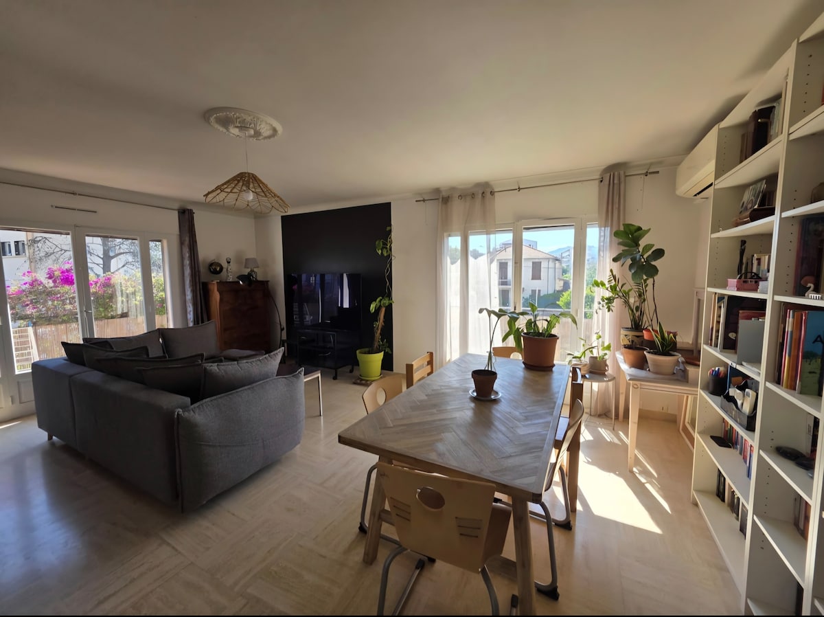 The spacious living area features a large gray sofa and a wooden dining table surrounded by light chairs. Natural light fills the room through large windows, highlighting potted plants. A black wall serves as the backdrop for a large television, complemented by simple decor.