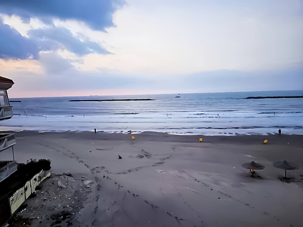 An expansive view of the ocean at twilight showcases gentle waves lapping against the sandy shore. The beach features scattered umbrellas and distant figures strolling along the water's edge. A soft gradient of pastel colors fills the sky, creating a serene coastal atmosphere.