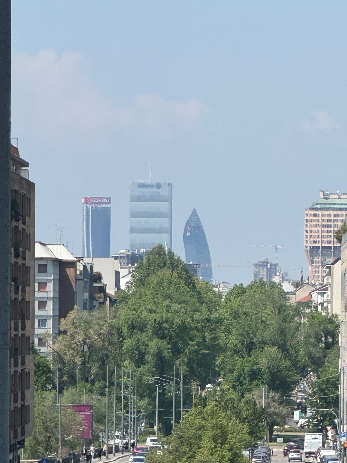 The skyline of Milan is visible, showcasing a variety of architectural styles. Several tall buildings are highlighted, including a prominently shaped structure. A row of trees runs along the roadway, adding greenery to the urban scene.