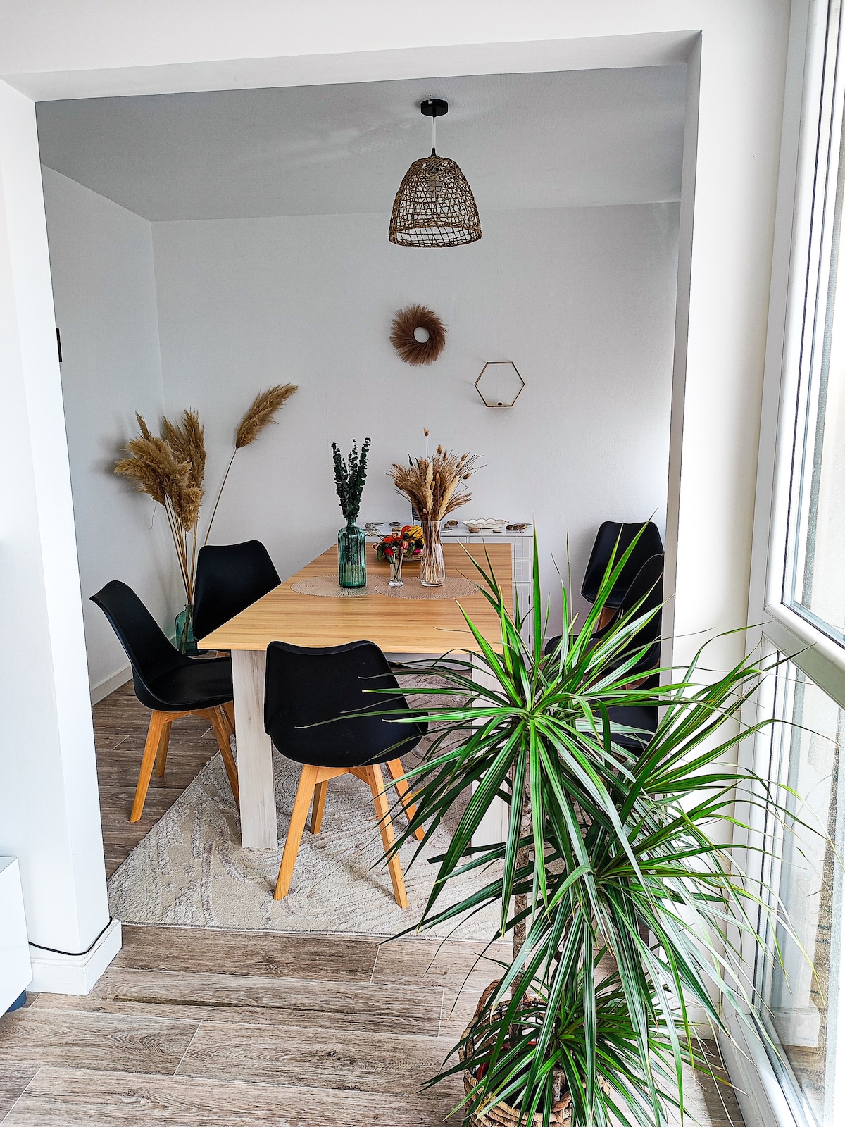 A dining area features a long wooden table surrounded by black chairs. Natural light enters through large windows, illuminating decorative dried plants in vases. A green potted plant is positioned nearby, adding a touch of greenery to the space. A simple light fixture hangs above the table.