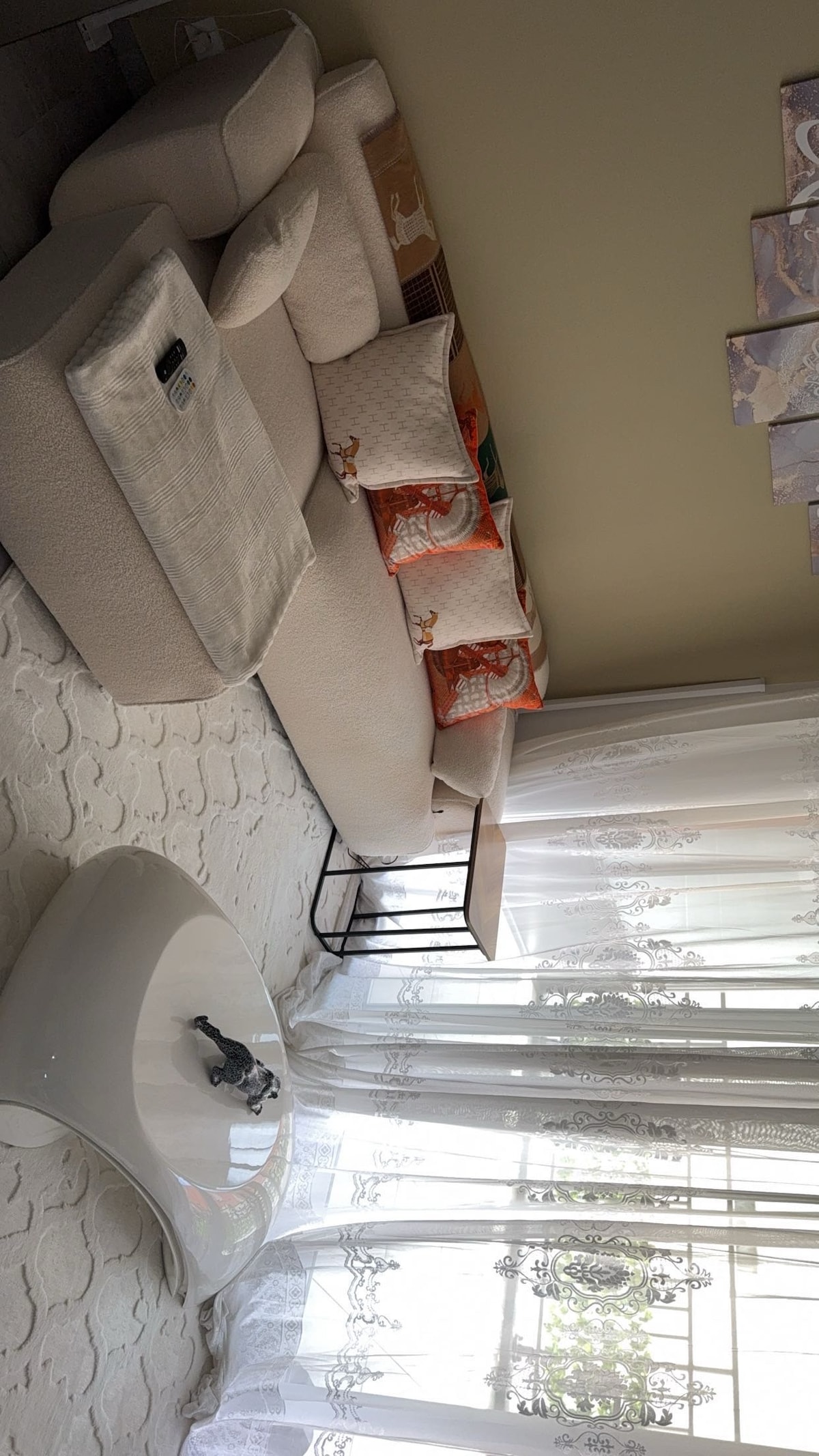 A light-colored sectional sofa is positioned against a wall, adorned with decorative pillows. A modern coffee table is placed in front, featuring a small decorative item. Large windows with sheer curtains allow natural light to filter into the inviting living area.