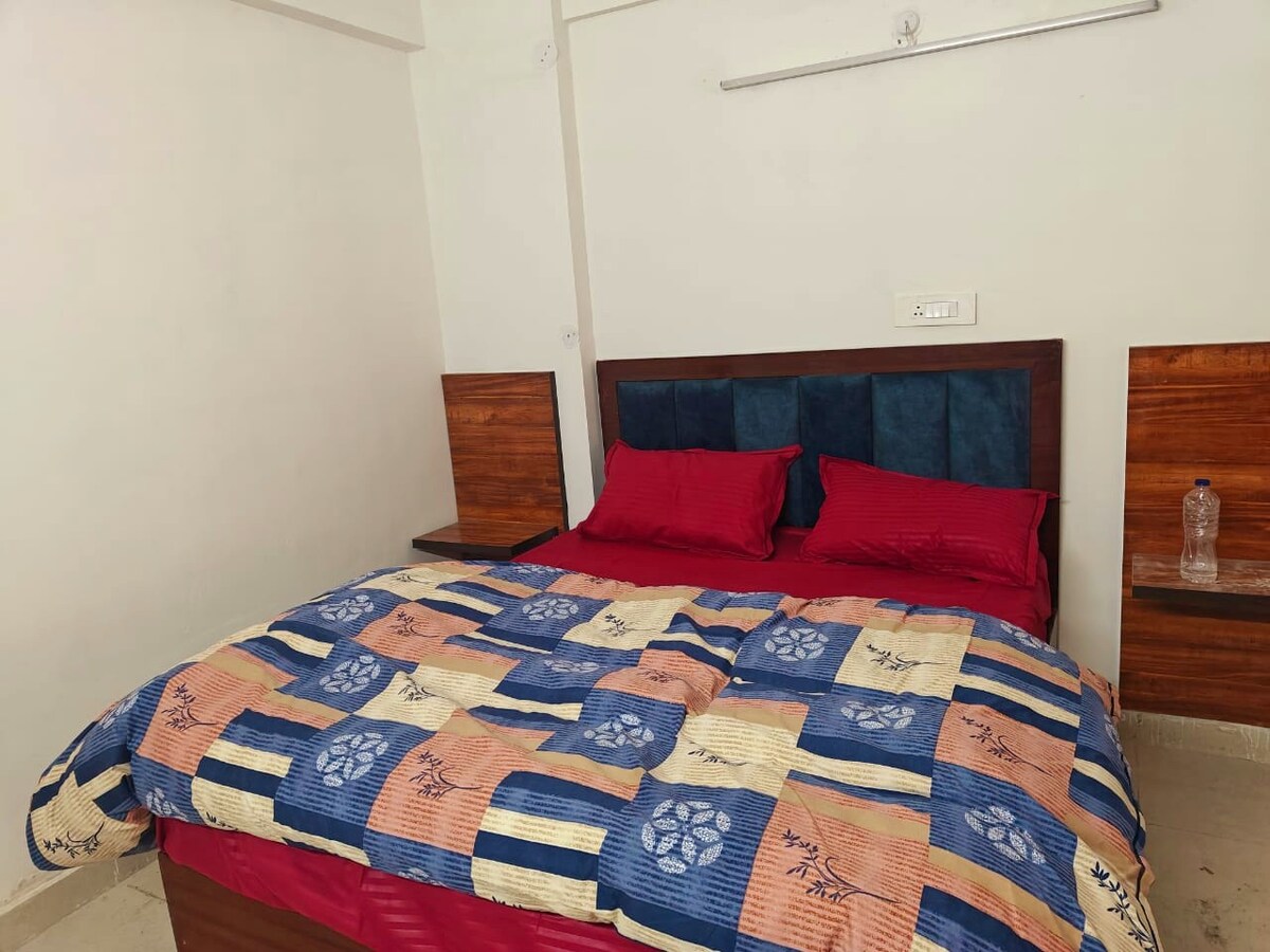 A cozy family room features a spacious bed covered with a patterned quilt in colorful hues. Two nightstands are placed on either side of the bed, and a water bottle is positioned on one nightstand. Light-colored flooring complements the overall decor.