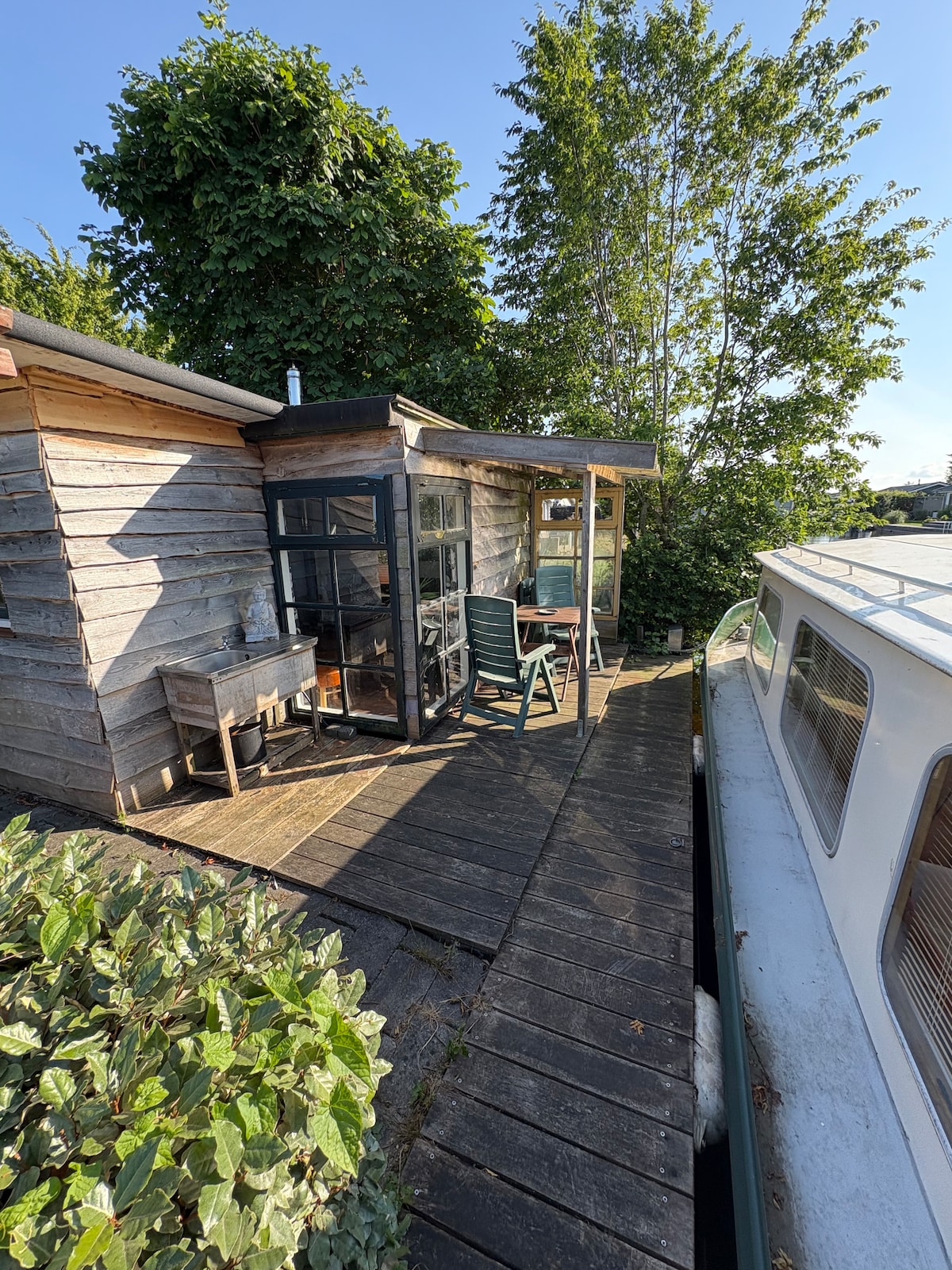 The image shows a wooden terrace adjacent to a house and a boat. A wooden structure with large windows provides natural light. There is a green chair positioned by the entrance, and a small table is visible. Lush greenery, including bushes and trees, surrounds the area.