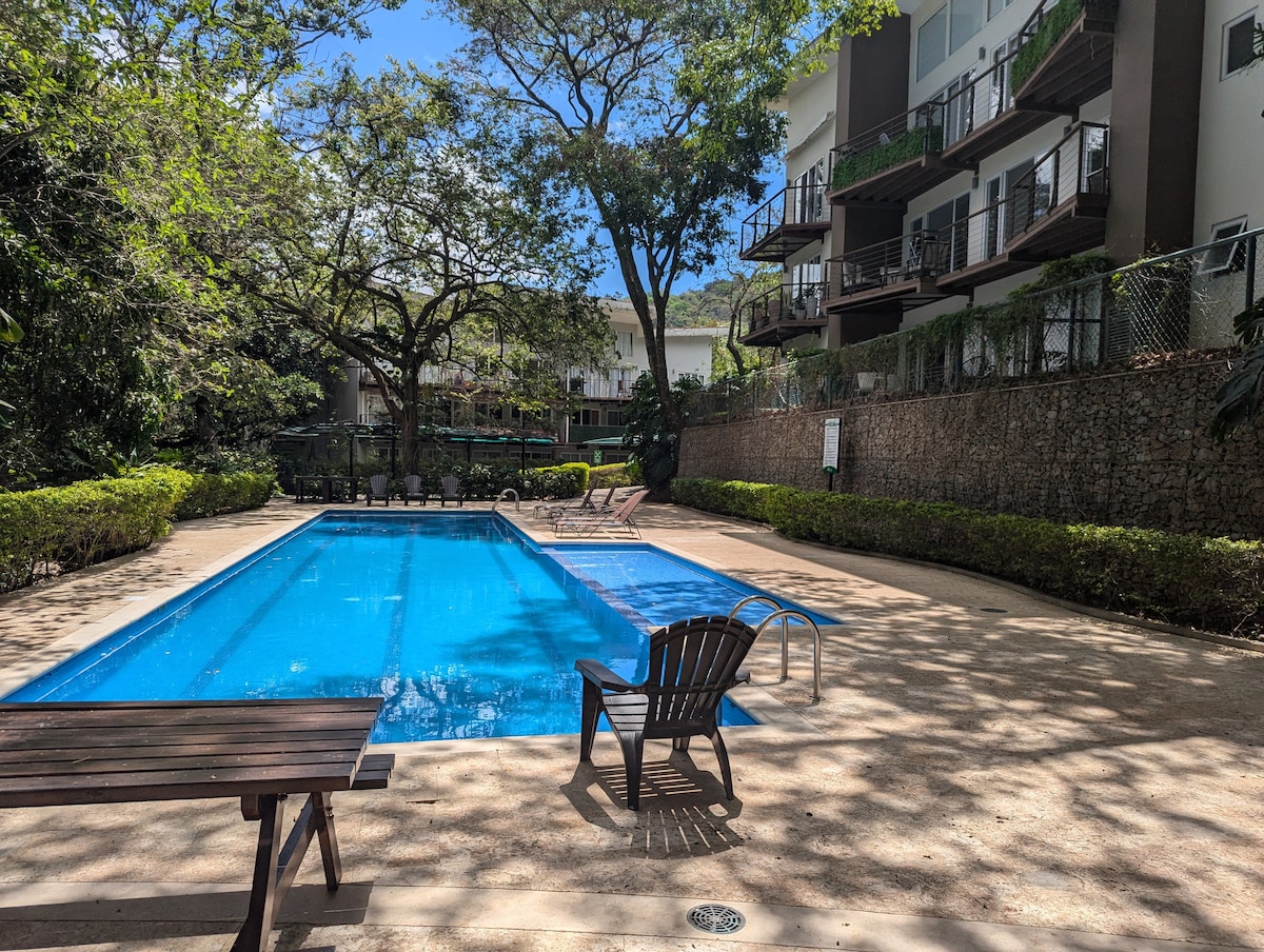 A serene outdoor pool area is surrounded by lush greenery and tall trees. The clear blue water reflects the sunlight, while wooden lounge chairs are positioned nearby. The building's balconies offer a view of the pool, providing a sense of tranquility and relaxation.
