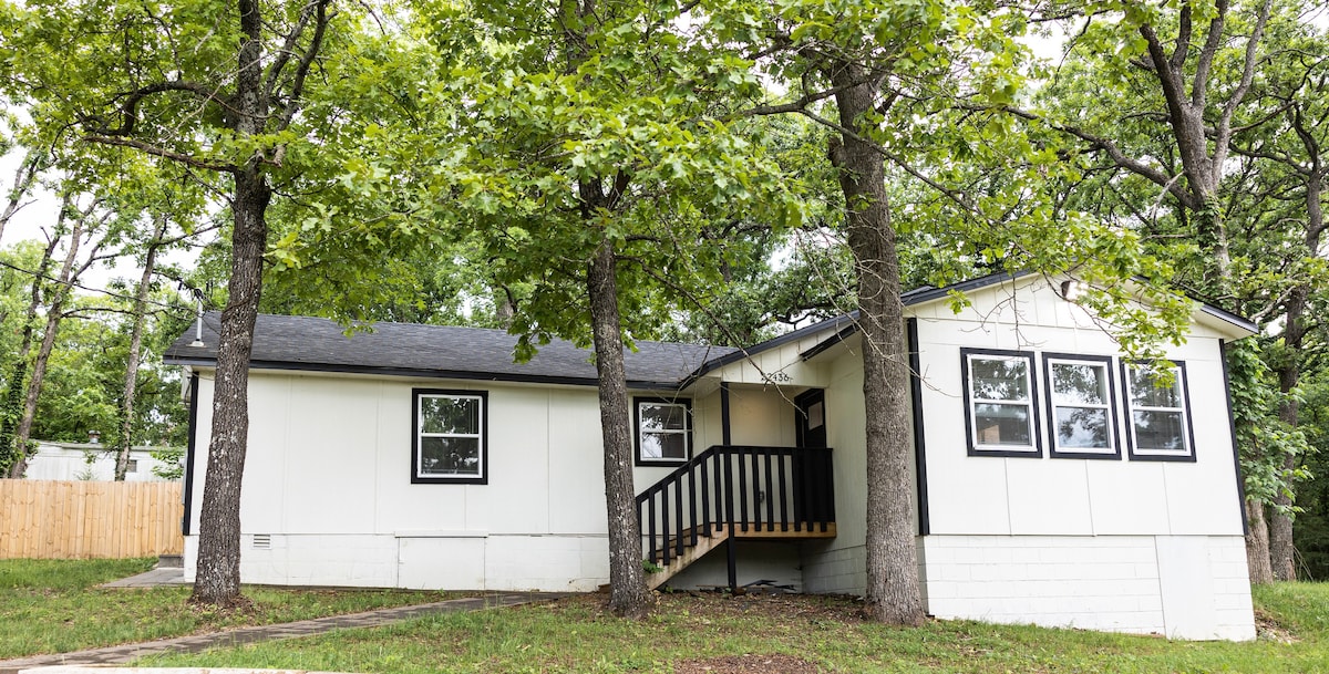 A welcoming family lake house is framed by tall trees, offering natural shade. The exterior features a mix of white and black elements, with a covered porch and black railing. Large windows allow for ample light, creating a bright and inviting facade.