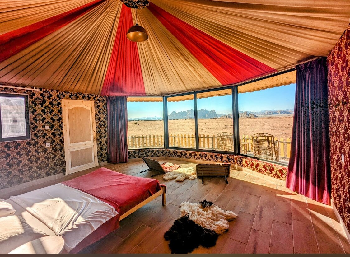 A spacious tent interior features large panoramic windows offering views of the desert landscape. The ceiling is adorned with colorful fabric draping, while a comfortable bed is positioned on natural wooden flooring. Chairs are placed near the windows, creating a cozy nook for relaxation.
