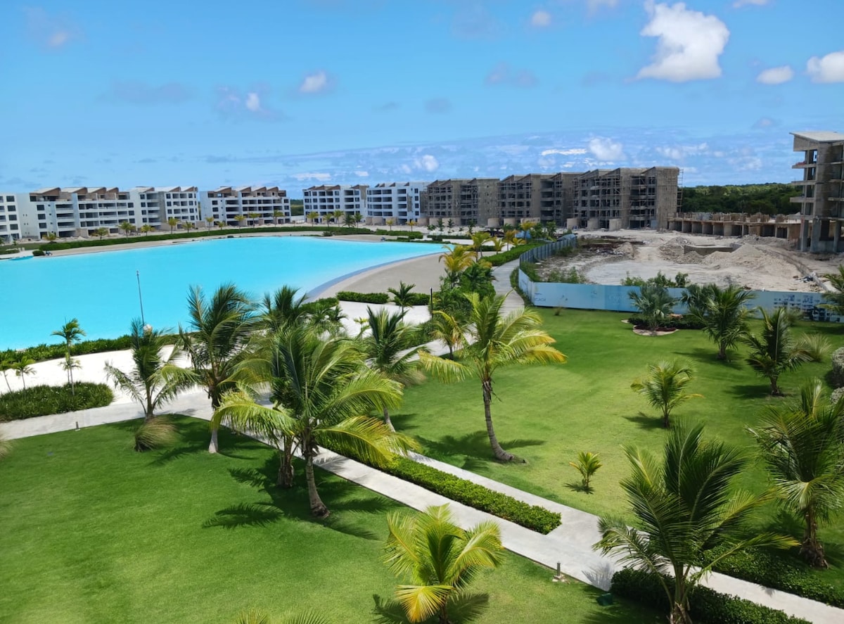 A panoramic view captures a vibrant green landscape featuring palm trees and manicured lawns. A large artificial lagoon with clear turquoise water is visible, surrounded by modern buildings in the background. The scene conveys a sense of openness and tranquility in a resort setting.