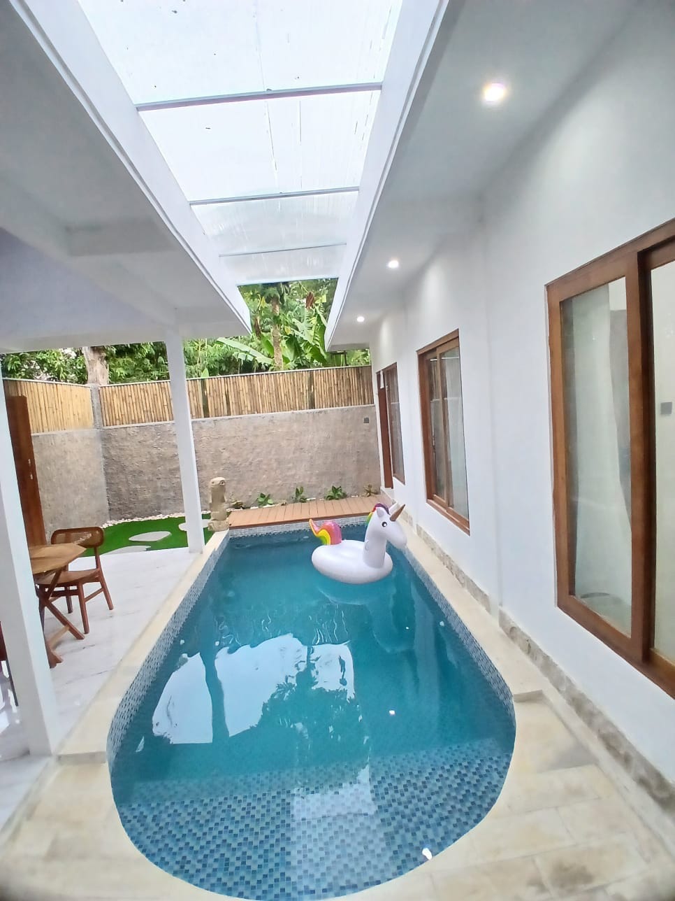 A private pool is situated in a well-lit area, surrounded by large windows and a clear roof. A colorful inflatable unicorn floats gently in the water. Wooden chairs and a small table are positioned nearby against a backdrop of natural greenery.