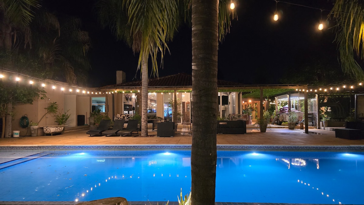 A serene pool area is illuminated by warm string lights, reflecting off the tranquil blue water. Lounge chairs are positioned nearby, and the surrounding greenery adds a natural touch. The modern building in the background features large windows that reveal a welcoming indoor space.