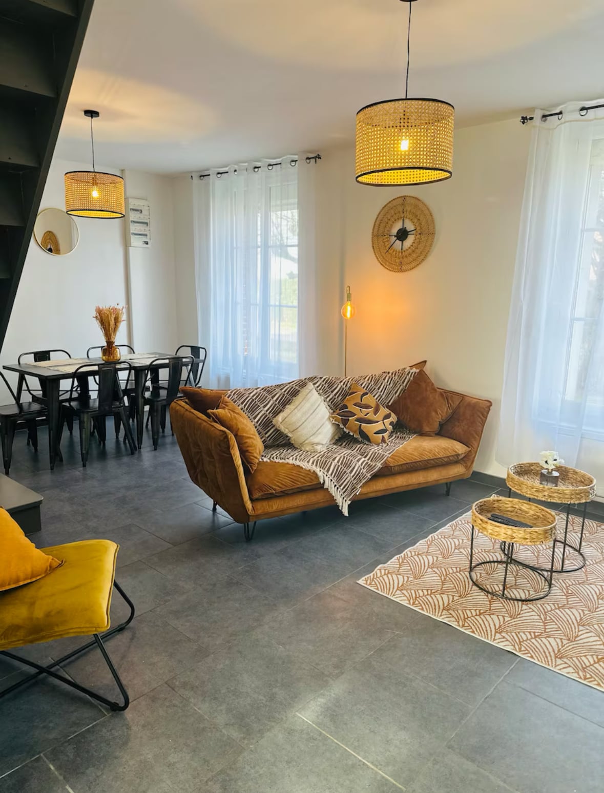 A cozy living area is presented with a brown sofa adorned with cushions and complemented by a patterned throw. Two small tables are arranged nearby, with a decorative item on one. The space is well-lit through large windows, with a dining table visible in the background.