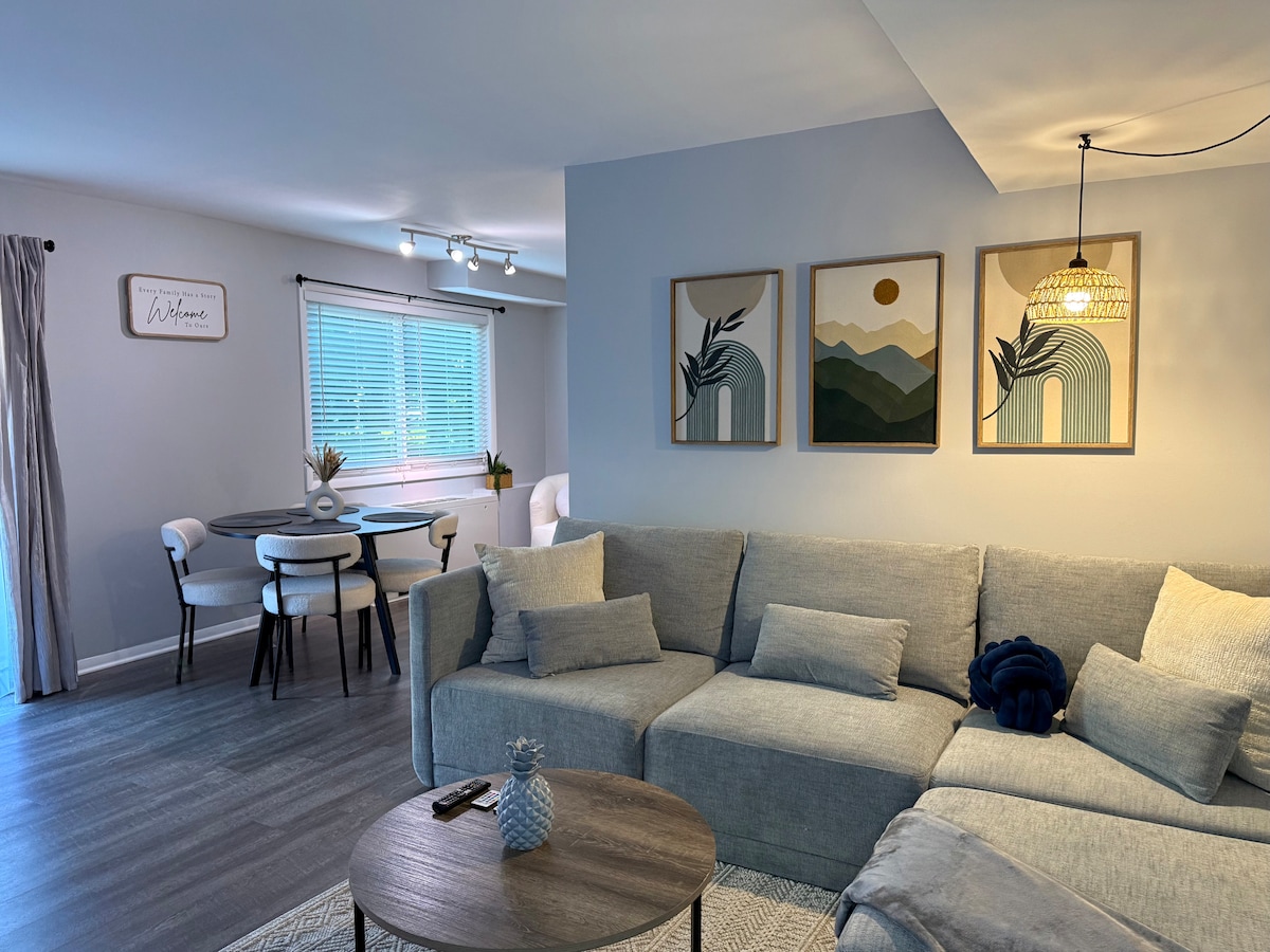 A cozy living space showcases a comfortable sectional sofa with neutral tones and textured pillows. A small wooden coffee table sits in front. Wall art featuring abstract landscapes is displayed above. A dining area with a round table and chairs is visible near a window with natural light.