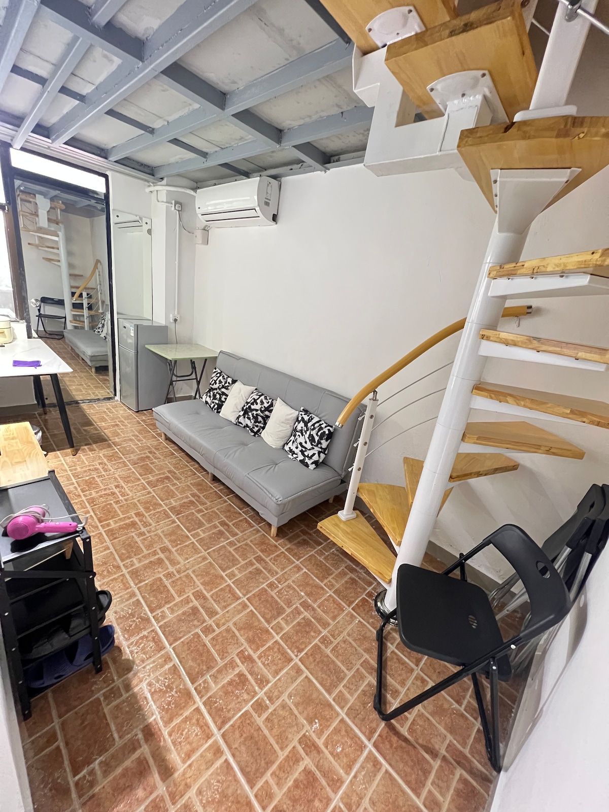 A spiral staircase leads to the upper level, while a gray sofa adorned with decorative pillows occupies the lower area. The floor features tiled flooring and a small table is positioned nearby. Air conditioning units are mounted on the wall, ensuring comfort in the space.