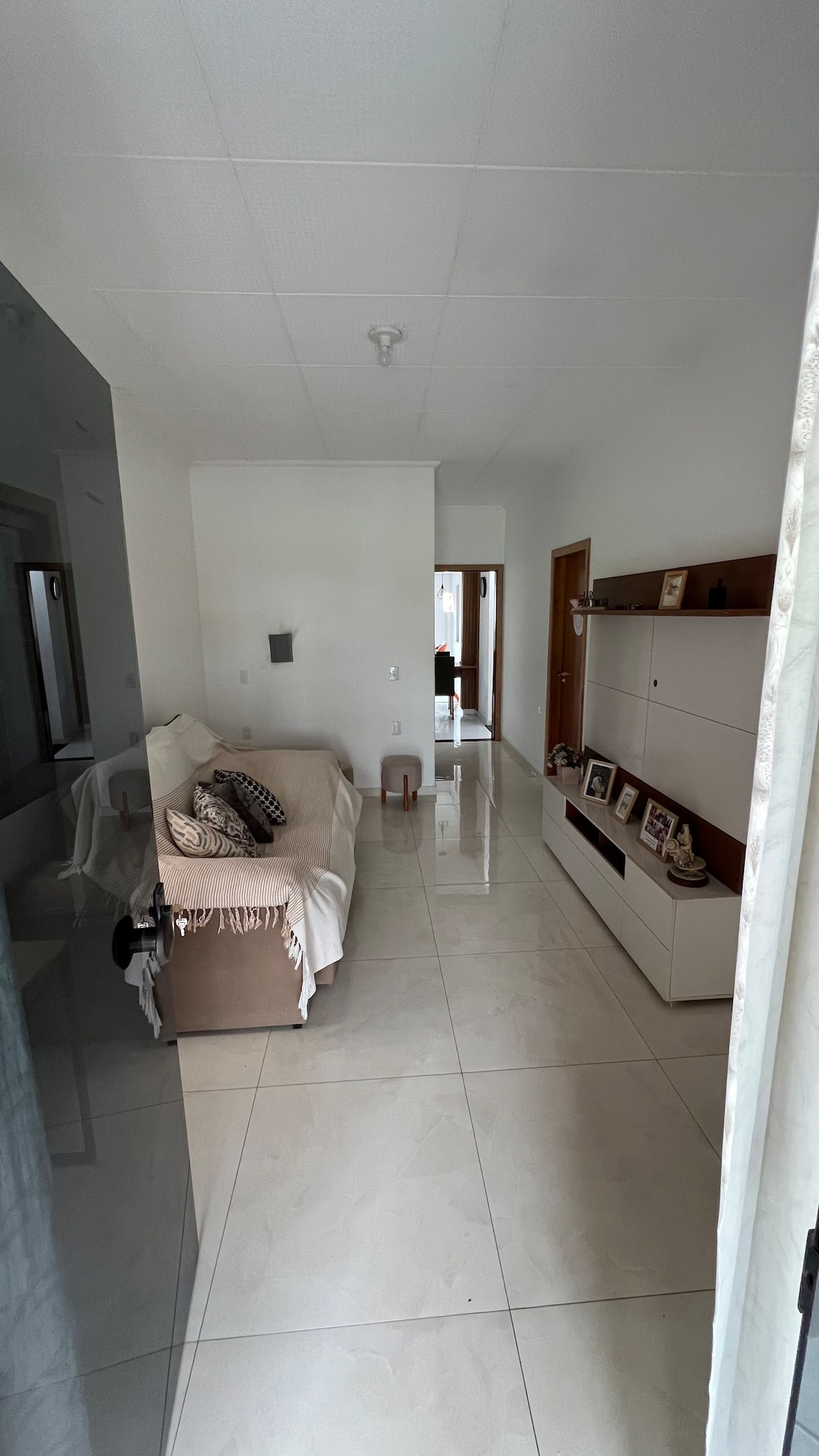 A well-lit hallway features shiny tiled flooring and white walls. A beige sofa is positioned along one side, complemented by decorative pillows. A modern console table holds picture frames and small items, while doorways are visible at the end of the hallway leading to other areas.