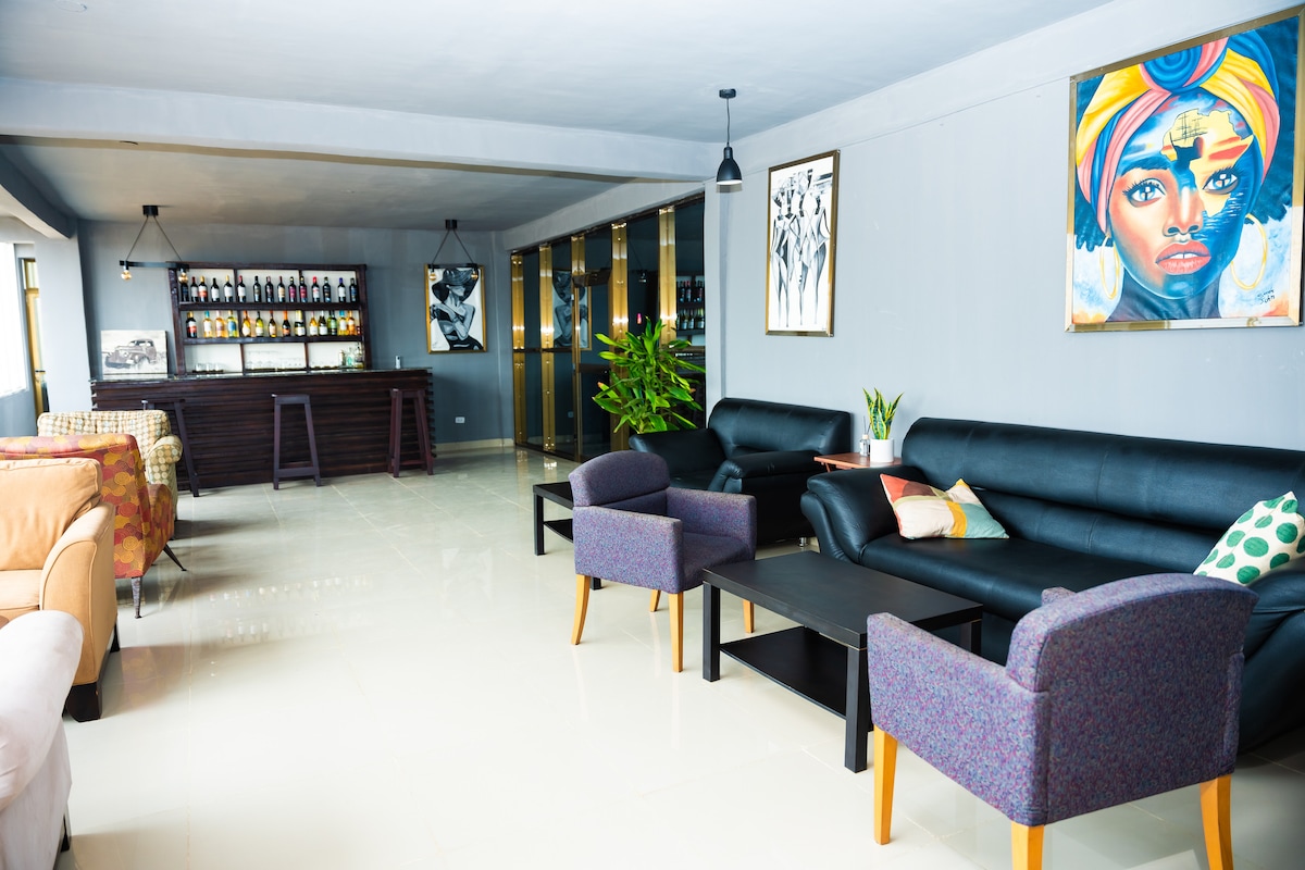 A spacious lounge area features a mix of comfortable seating, including black leather sofas and accent chairs. A sleek bar unit is positioned against the wall, showcasing a selection of drinks. Art pieces adorn the walls, complemented by natural light entering through large windows.