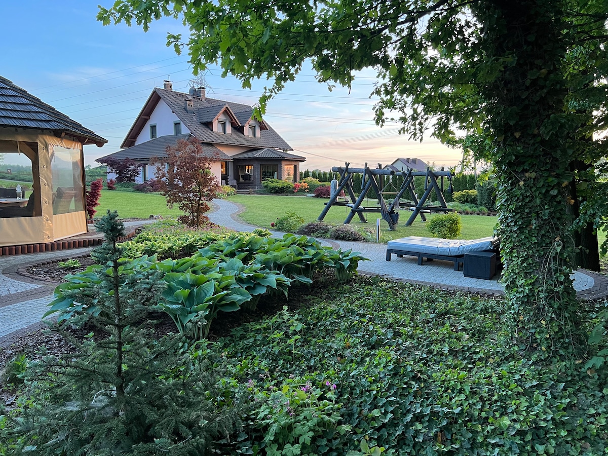 A spacious garden area is highlighted by lush greenery and vibrant foliage. A pathway leads to a welcoming house in the background. Outdoor seating and shaded areas are visible, along with a playground structure, providing a serene environment for relaxation and play.