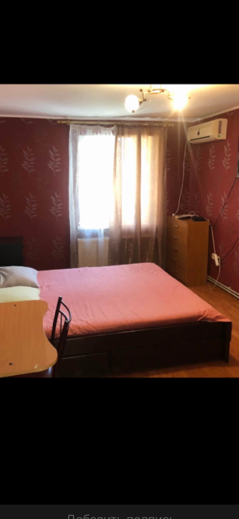 A comfortable sleeping area features a spacious bed with a soft pink coverlet. Natural light enters through a window framed by sheer curtains. A wooden dresser stands nearby, while a small table and chair provide additional furnishings against the patterned red wall.