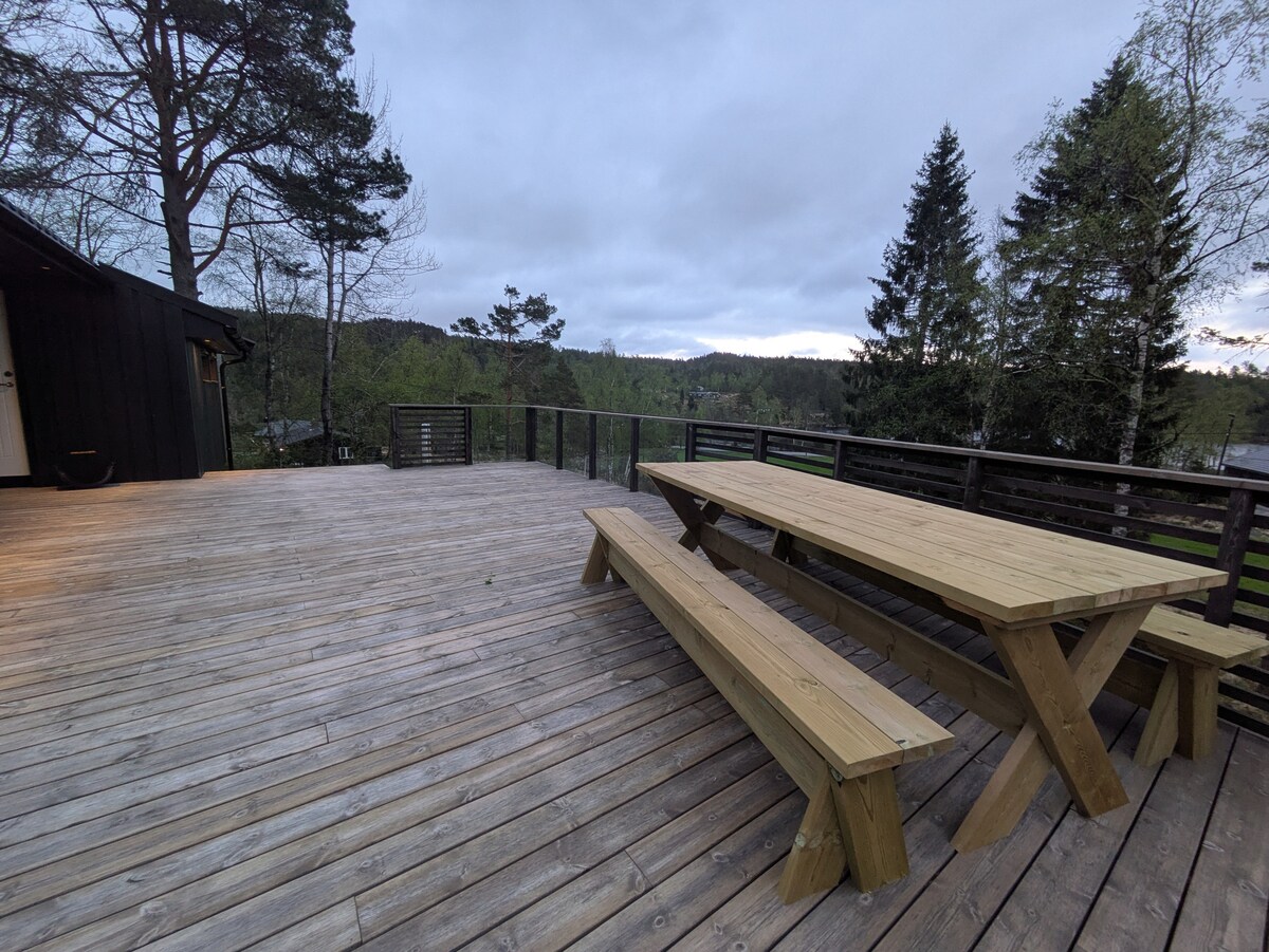 A spacious wooden deck features a large picnic-style table and benches, designed for outdoor dining. Surrounded by trees, the area provides views of the natural landscape, with a soft twilight glow enhancing the serene atmosphere.