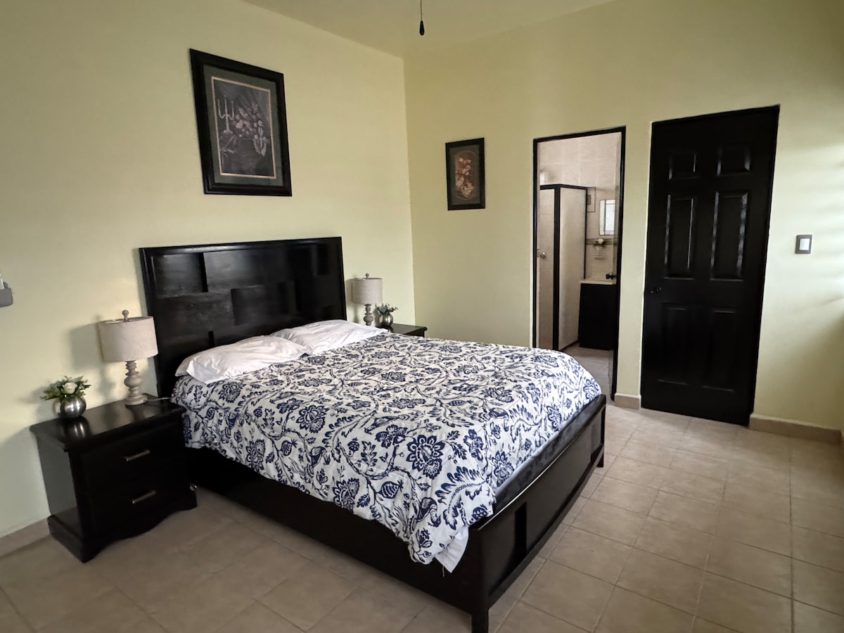 A spacious bedroom features a dark wood bed frame with a light blue and white patterned comforter. Nightstands with lamps are positioned on either side of the bed. The room is illuminated by natural light, complemented by light green walls and framed artwork.