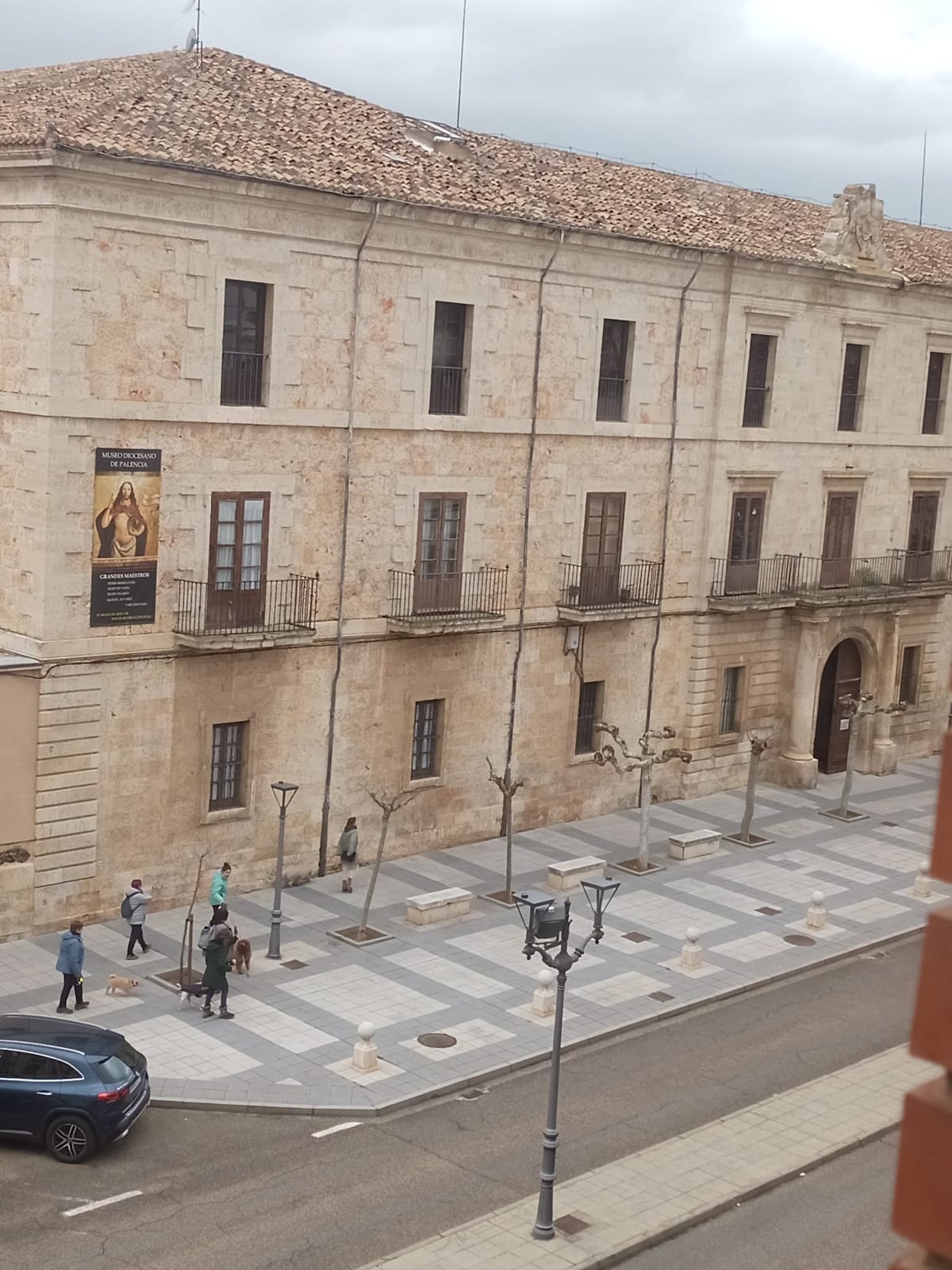 The image showcases a historic building with a stone facade and a tiled roof. Pedestrians can be seen walking along a patterned street lined with trees. Street lamps are visible, contributing to the urban landscape.