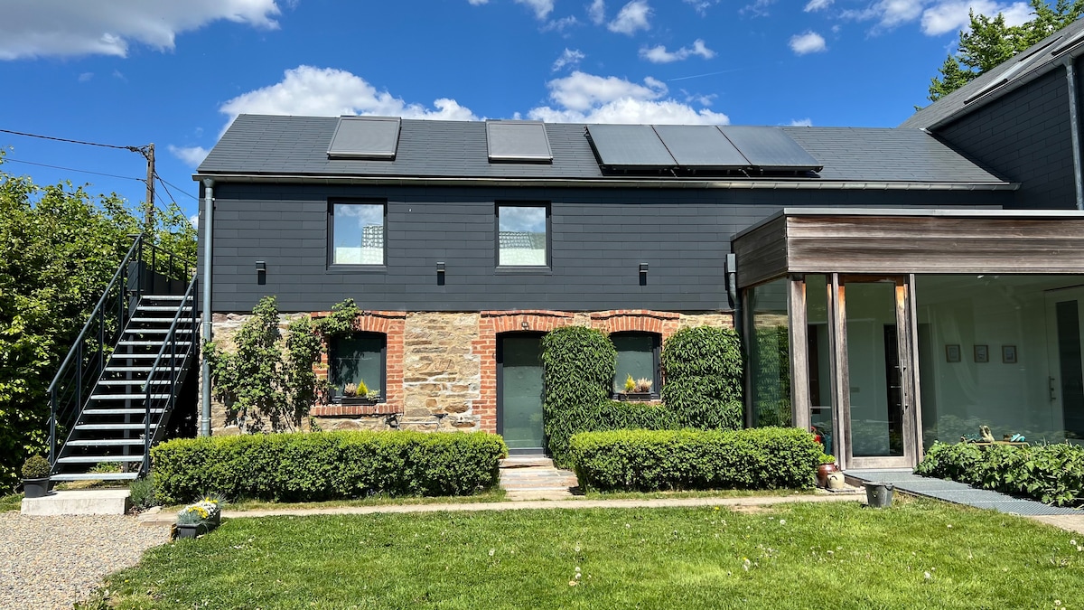 A contemporary lodge exterior is presented, with solar panels on the roof and a combination of stone and dark siding. Greenery frames the entrance, featuring a landscaped pathway. An outdoor staircase leads to the upper level, while large windows invite natural light into the space.