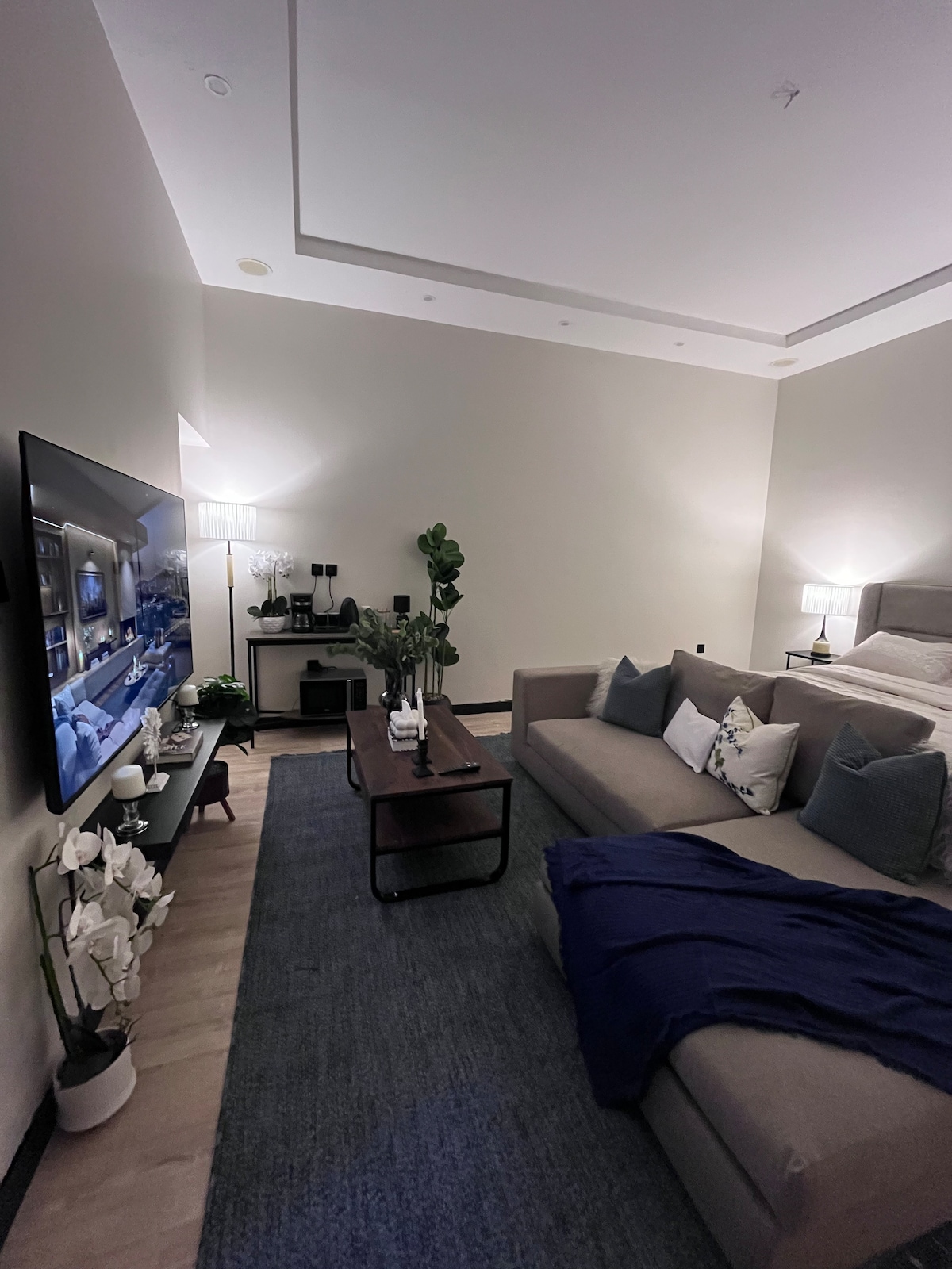A modern living area features a comfortable sectional sofa adorned with decorative pillows. A sleek coffee table is positioned in front, accompanied by decorative items. A wall-mounted television and a cozy bed in the background create a welcoming and functional space, illuminated by soft lighting.