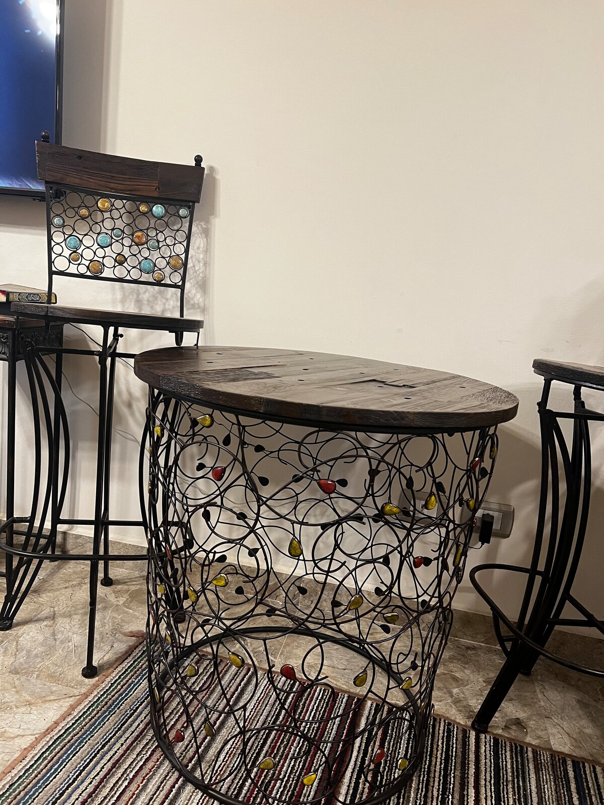 A uniquely designed table features an intricately woven metal base with colored accents. The tabletop is made of wood, adding a natural element. Nearby, a high-backed chair with similar wire detailing complements the table, set against a soft-textured rug.