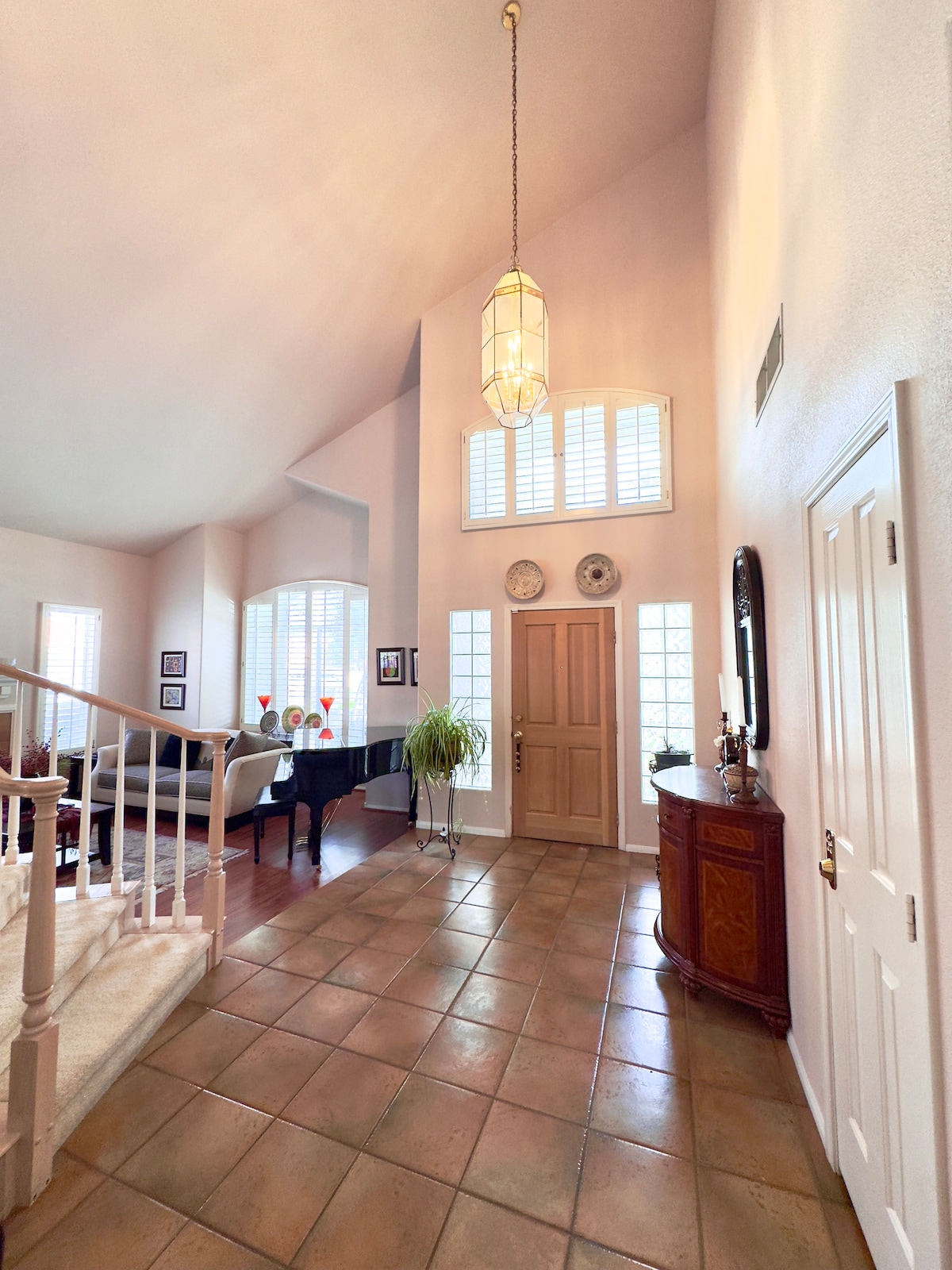 An inviting entryway features a high ceiling and rich wood flooring. Large windows allow natural light to illuminate the space. A grand piano is visible in the background, complemented by decorative wall art and a potted plant. The area is defined by tile flooring and elegant fixtures.