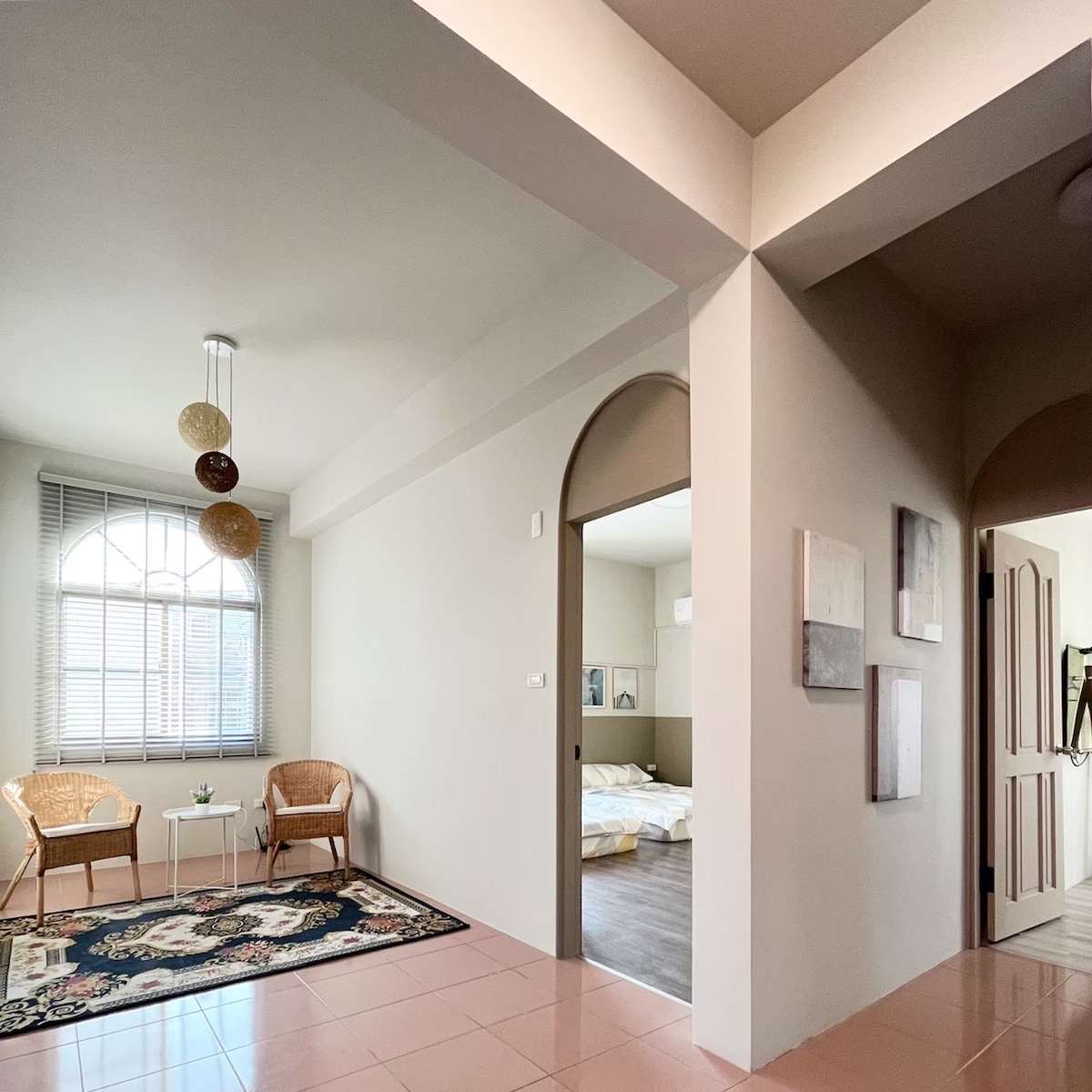 An open space is created, featuring two rattan chairs arranged beside a small table on a patterned rug. Arched doorways lead to additional areas, where a well-lit sleeping area with a spacious bed can be seen in the background.