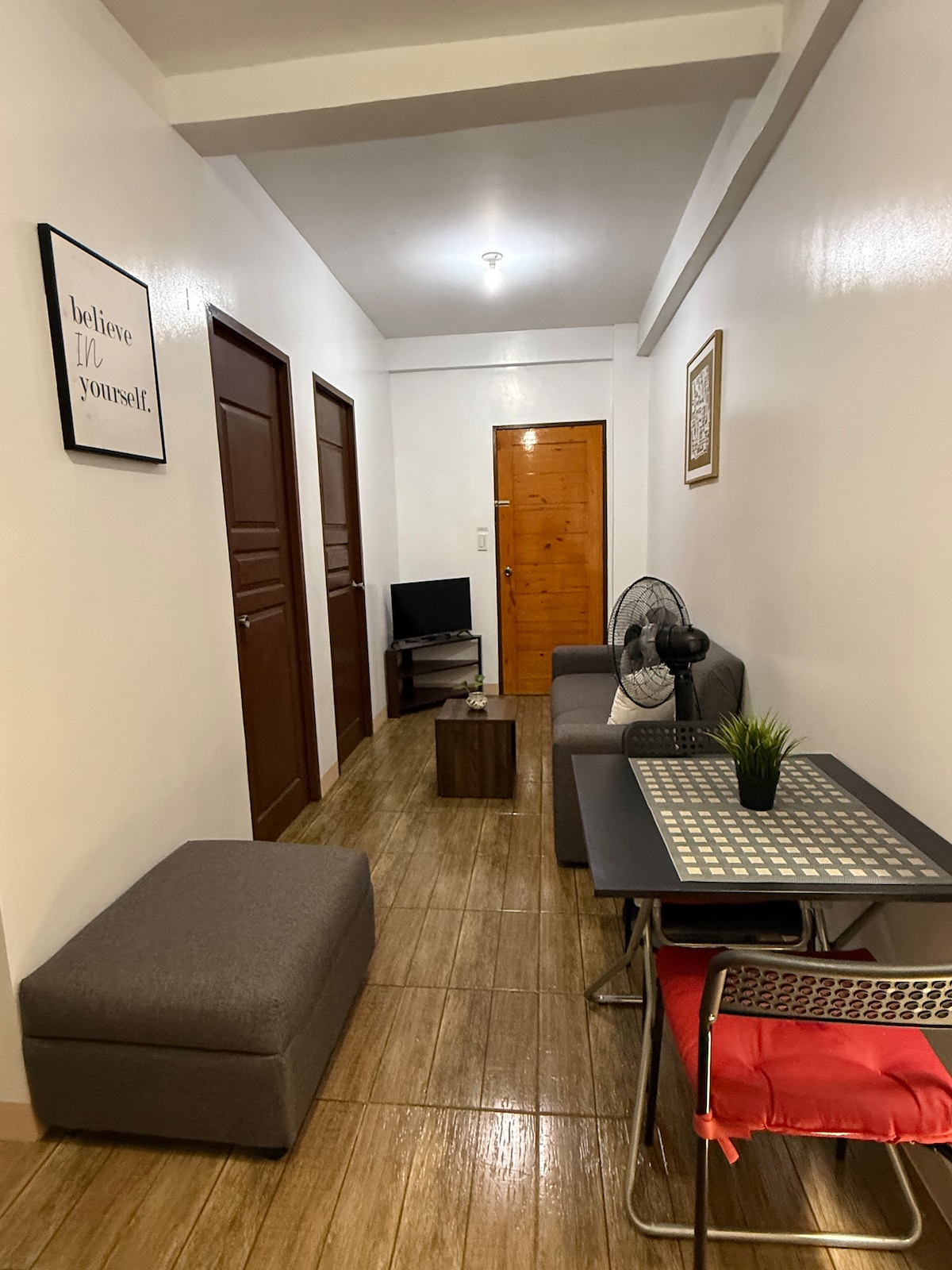 A narrow hallway is depicted, featuring wooden doors leading to rooms on either side. A small seating area is occupied by a dark sofa and an ottoman, while a compact table and chairs are positioned nearby. A television is mounted on a small cabinet, and a fan is visible.