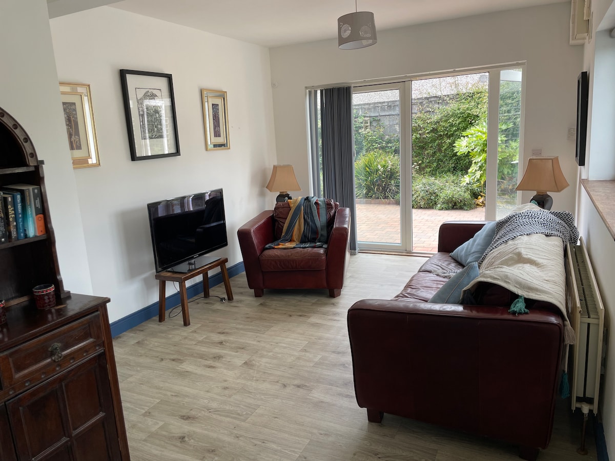 A spacious living area features two brown leather sofas, each adorned with throw blankets. A television stands on a console opposite, while large glass doors provide views of the outdoor space. Neutral tones and plants create a serene ambiance.