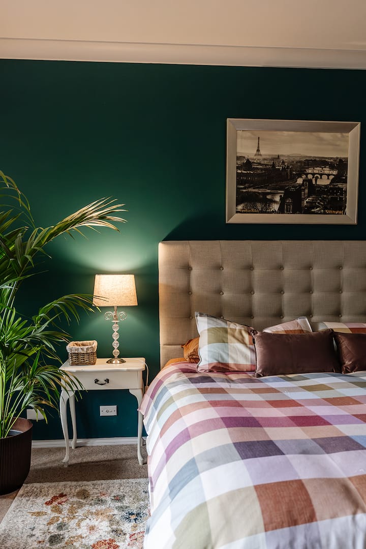 Cosy king bedroom with deep teal feature wall, upholstered linen bedhead, plaid bedding in warm tones, and a bedside lamp creating soft ambient lighting.