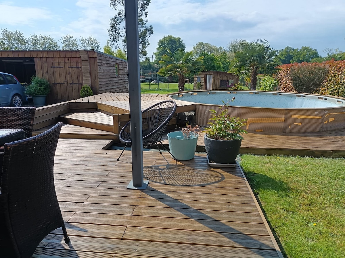 A wooden deck surrounds a rounded pool, highlighted by lush greenery and potted plants. Comfortable outdoor seating is positioned near the pool, and well-maintained grass complements the space. The area is brightened by natural light, showcasing the inviting outdoor setting.