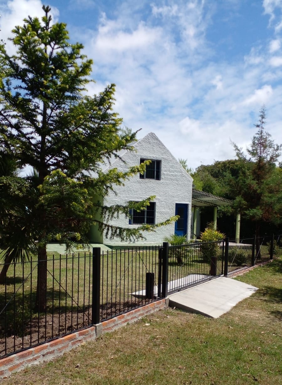 A two-story cabin is depicted, featuring textured white walls and blue window frames. A covered porch extends along the front. Surrounding greenery includes coniferous trees, and a fenced yard is visible, providing an inviting outdoor space for guests.