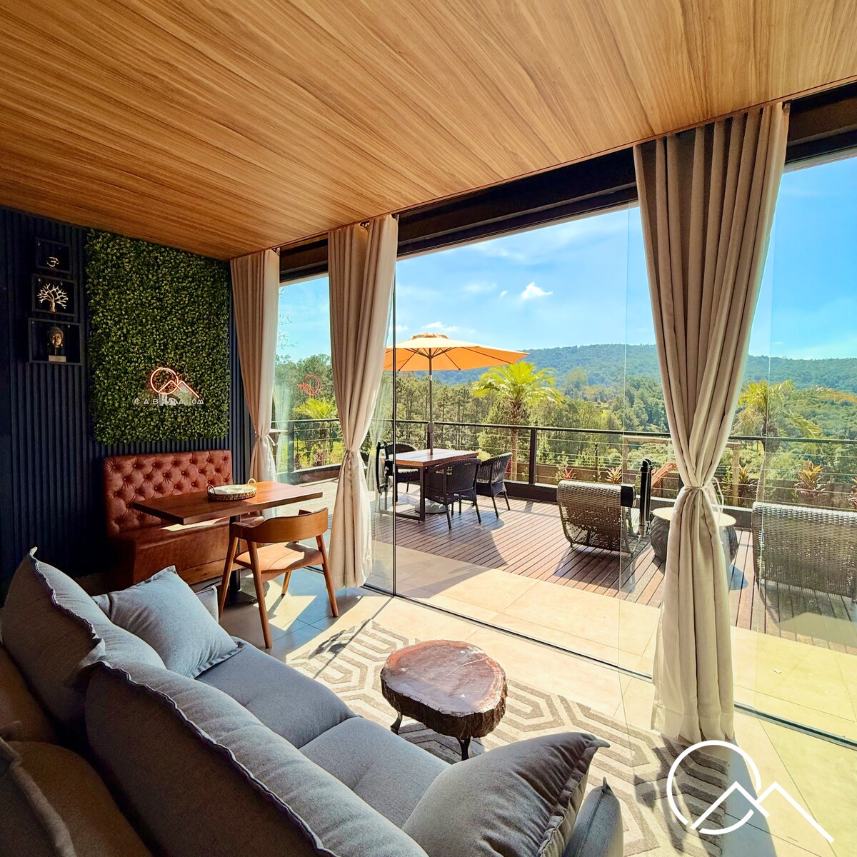 A bright and spacious living area is seen through large glass doors, providing a view of lush mountains in the distance. Comfortable sofas are arranged invitingly, alongside a wooden table. An outdoor patio is visible, featuring seating and an umbrella for shade.
