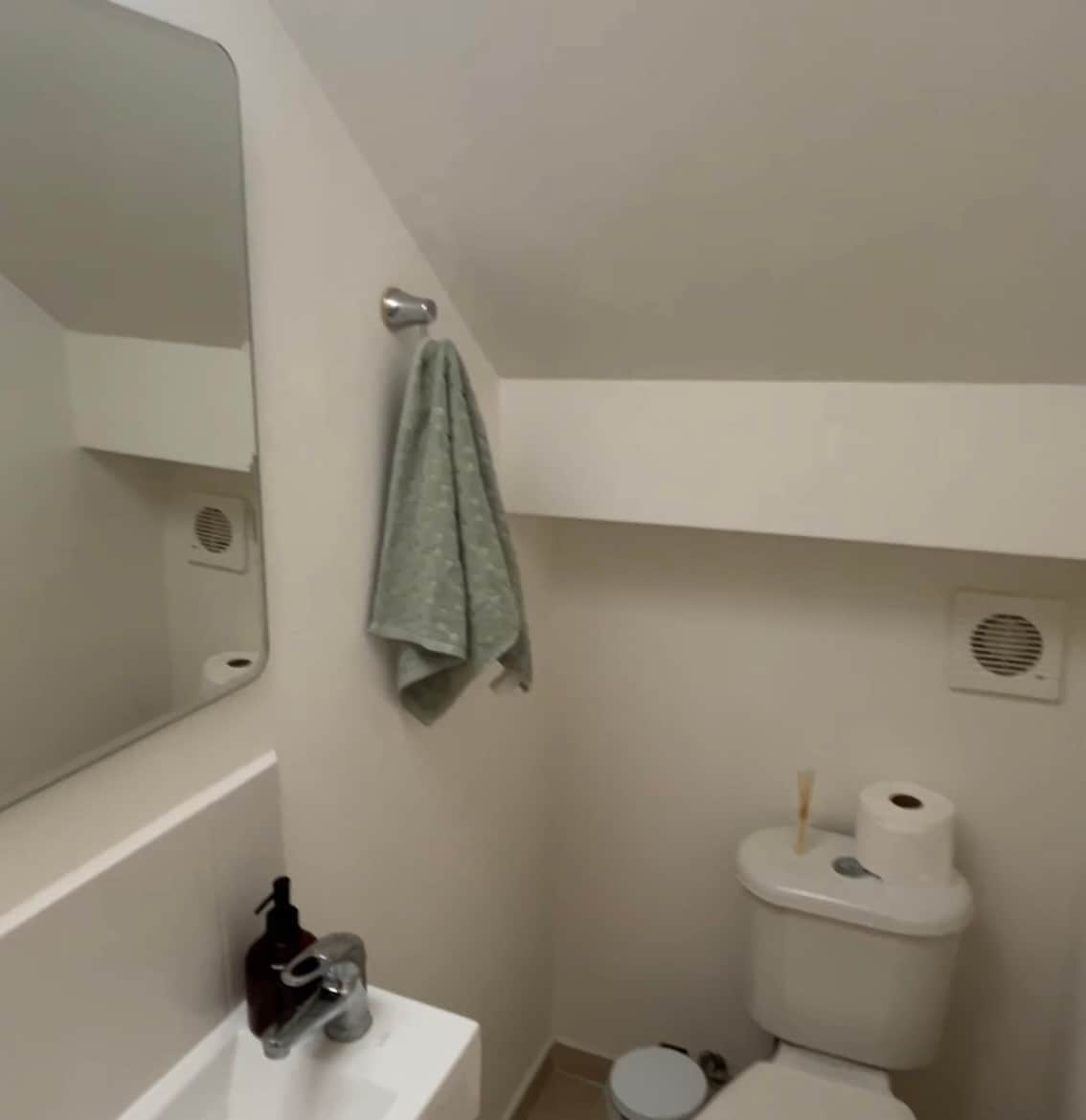 A compact bathroom is presented, featuring a wall-mounted mirror above a sink. A hand towel hangs on a silver hook nearby. A toilet is positioned against the wall, accompanied by a roll of toilet paper. Ventilation grilles can also be seen.