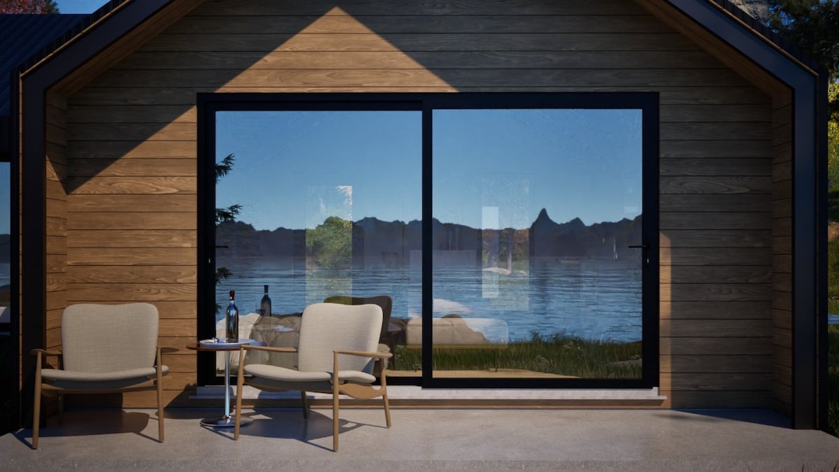 A cabin's exterior is shown with large glass doors offering views of a serene lake and mountains. Two light-colored chairs and a small table are positioned on a patio, creating an inviting space to relax and enjoy the scenery.