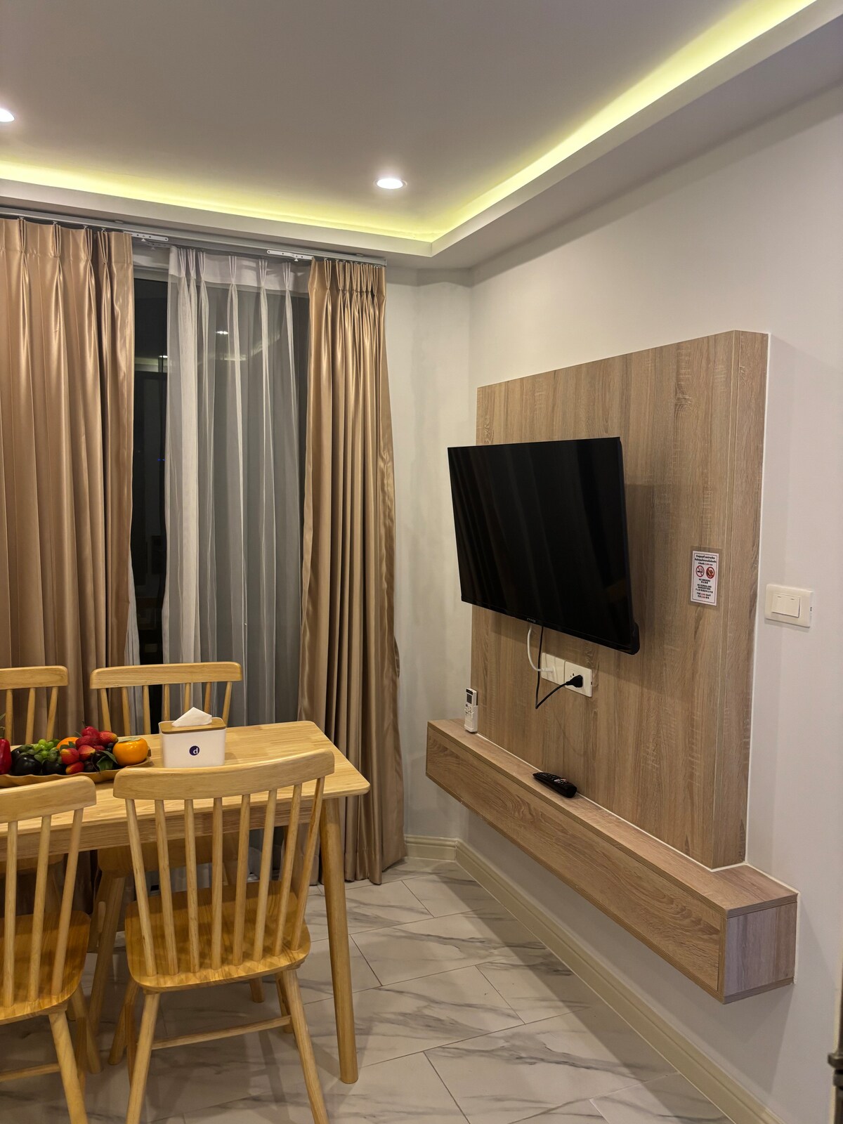 A dining area features a wooden table surrounded by four light-colored chairs. A flat-screen television is mounted on the wall, with a small shelf beneath. Floor-to-ceiling sheer curtains allow natural light to enter, complementing the neutral color scheme of the room.