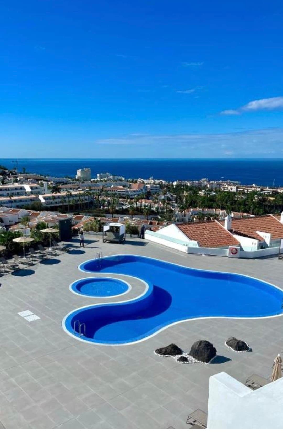 An expansive swimming pool is featured, shaped organically and surrounded by a spacious tiled deck. The view encompasses a vibrant coastal landscape with buildings and greenery, as well as the sea stretching into the distance under a clear blue sky.